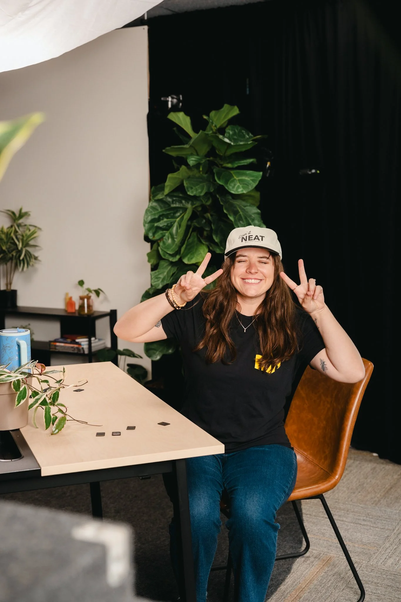A young woman sitting in a brown chair at a table with plants, smiling, making peace signs with both hands, wearing a white cap that says NEAT, a black t-shirt, and blue jeans, in a room with green plants and black curtains.