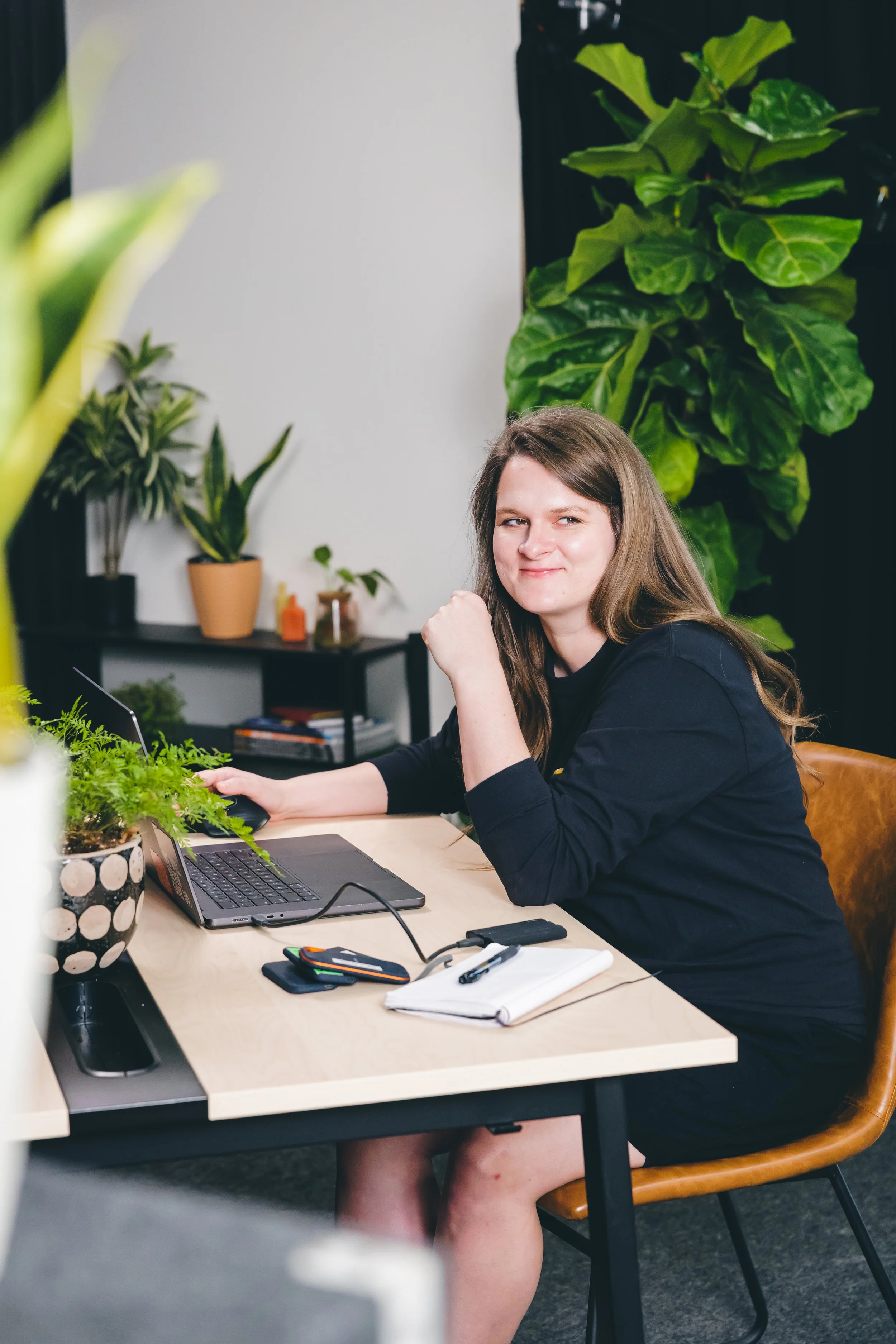 A woman sits at a desk with a laptop, surrounded by green plants in a modern office or home workspace.
