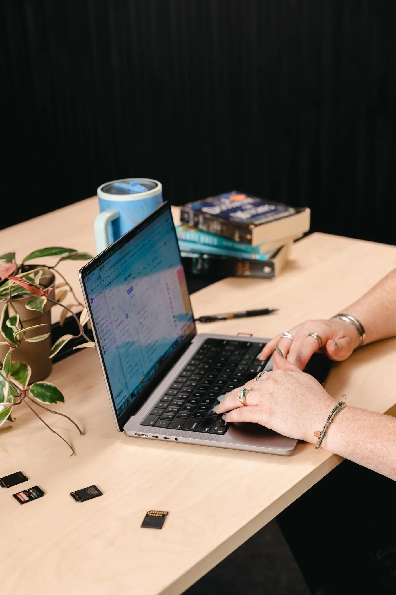 Person typing on a laptop at a wooden desk, with books, a mug, plant, and SD cards around.