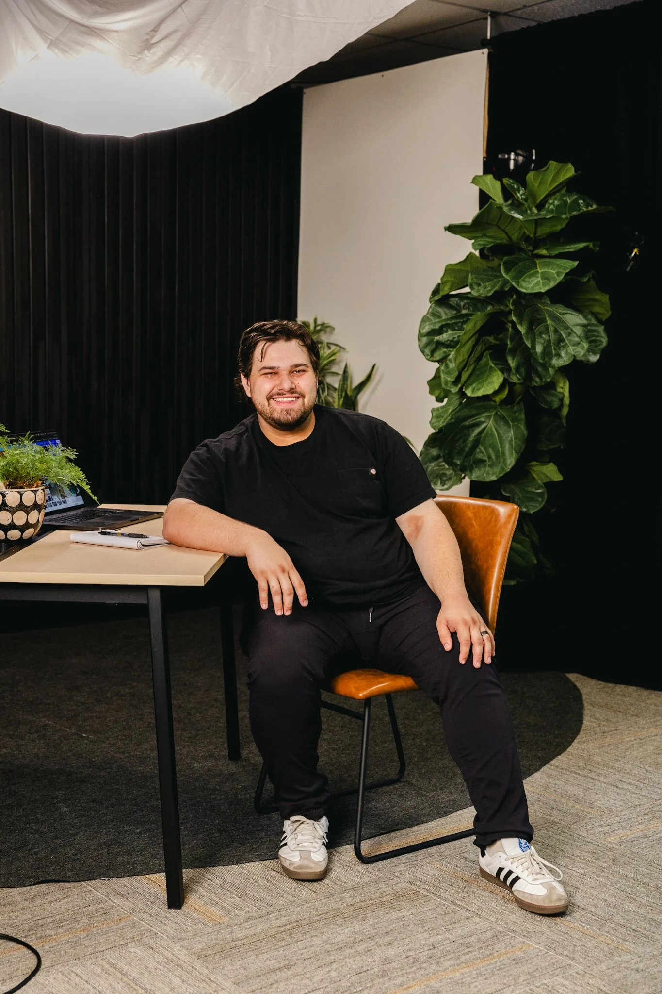 A man in all black with Adidas shoes smiling for the camera. He is sitting at a desk with a laptop, plants, and a book nearby. There are plants behind him and he is in a tan chair