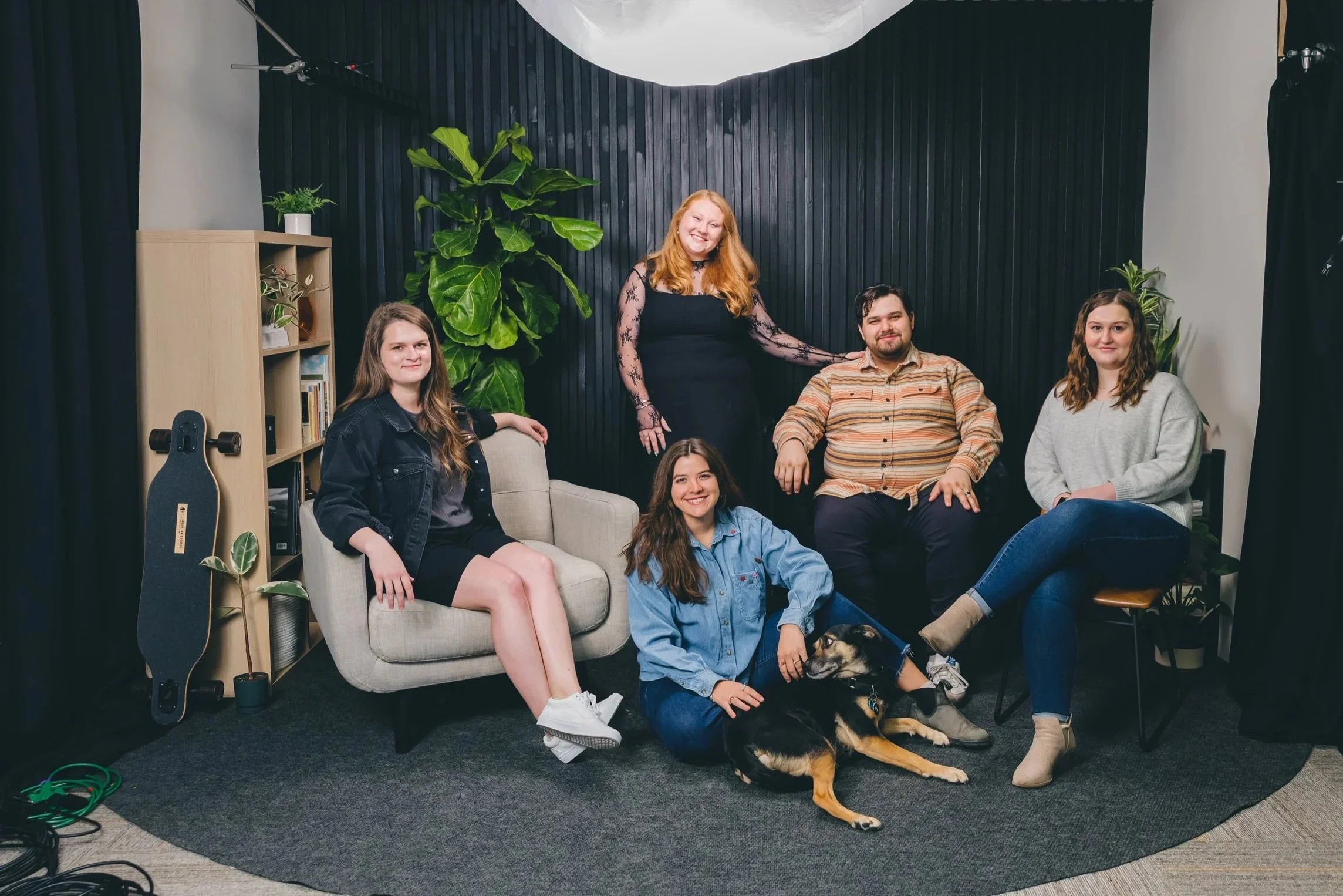 Group of five young adults and a dog in a studio with dark walls, a large green plant, bookshelves, and skateboards, smiling for the photo.