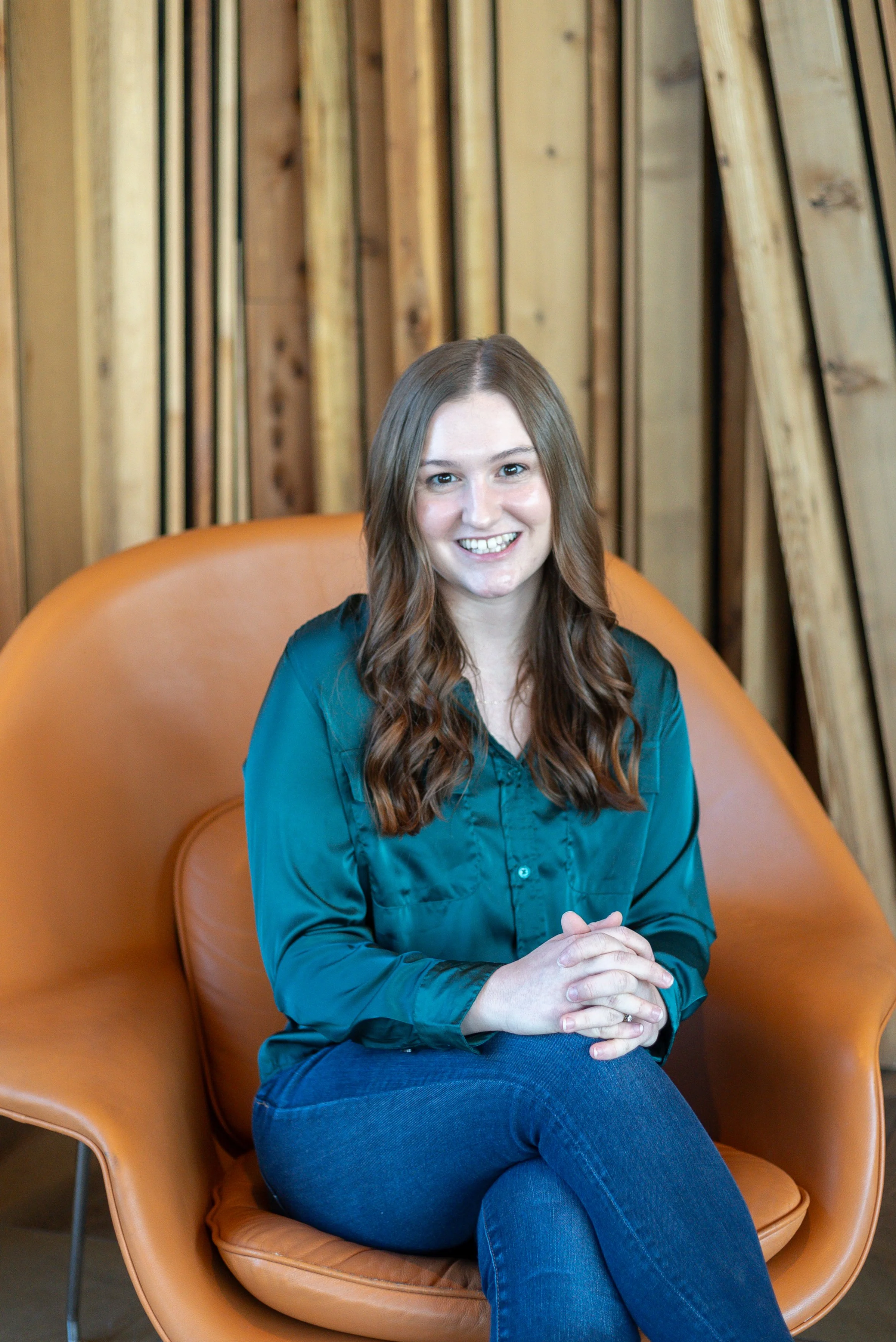 A young woman with shoulder-length black hair smiling brightly, wearing a blue top, standing among green plants in a greenhouse or garden.