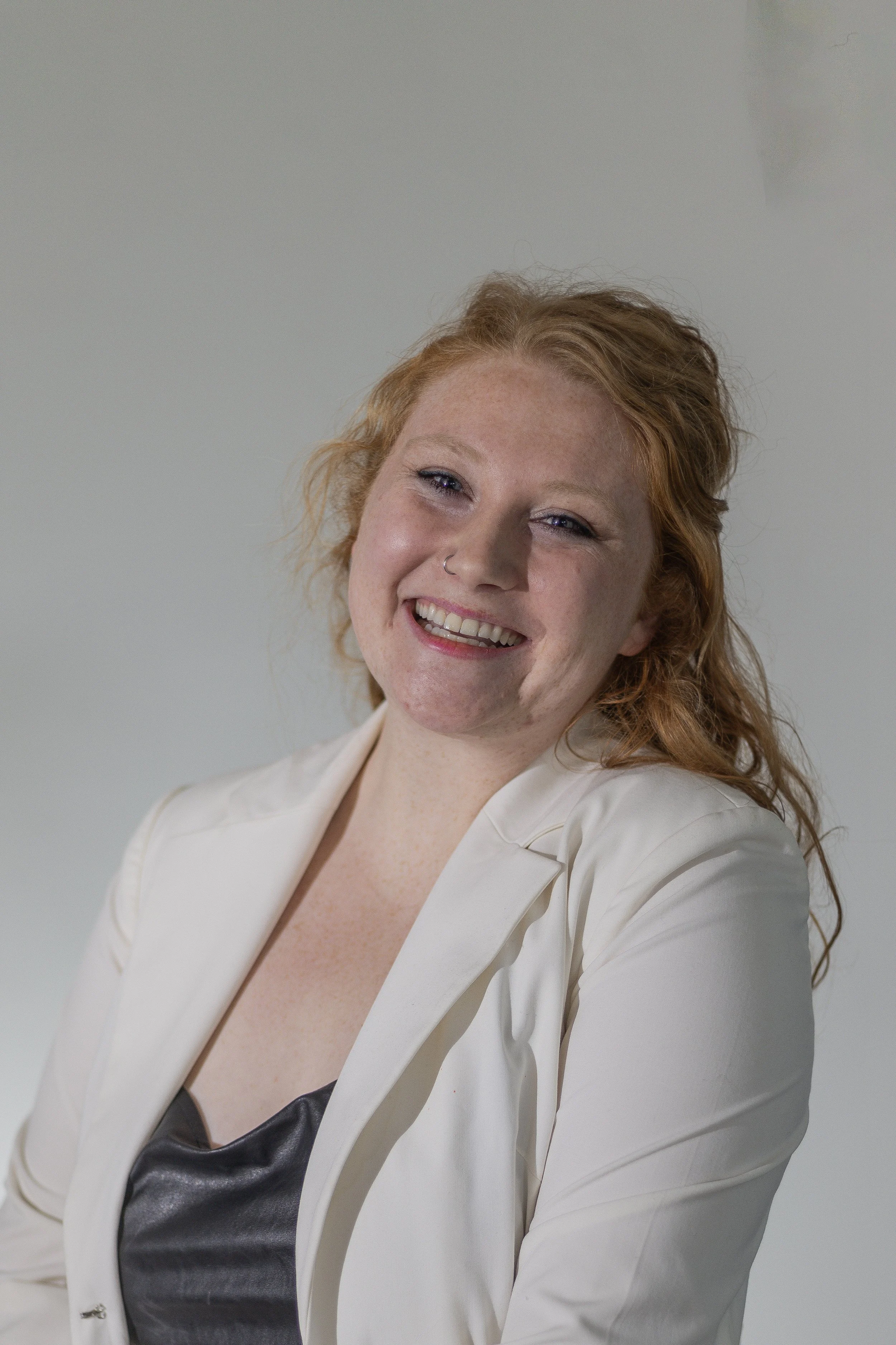 A woman with curly red hair, fair skin, and a nose piercing smiling, wearing a white blazer over a black top, against a plain light background.