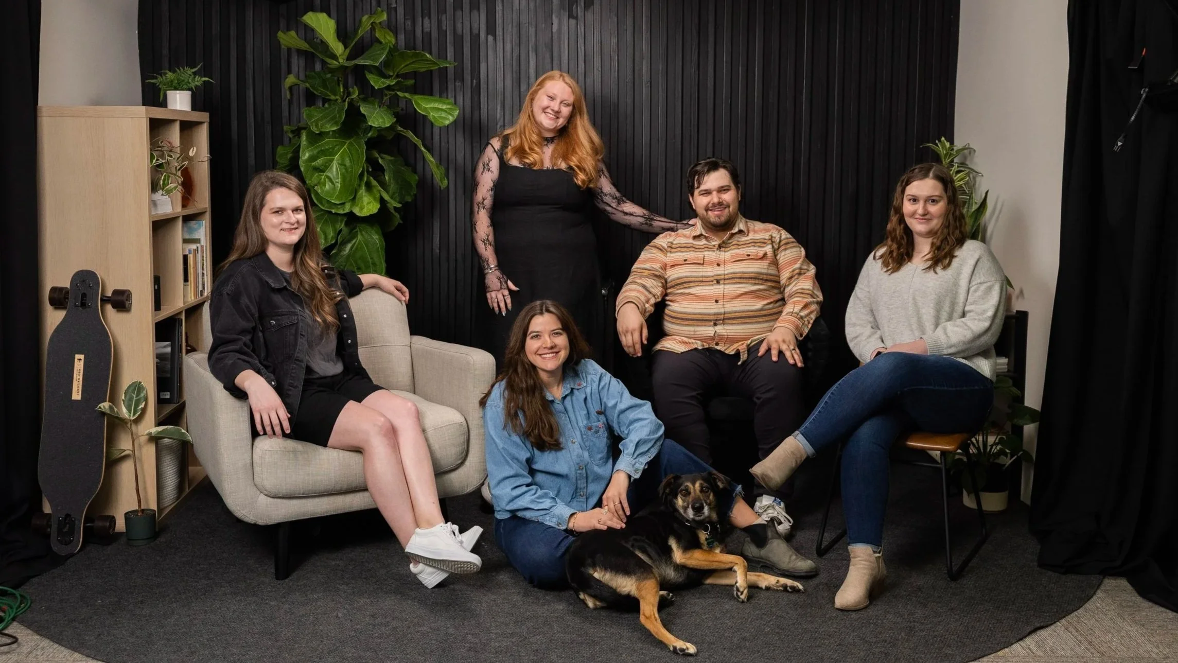 Group of five people with a dog in a cozy indoor setting with black wall, plants, and furniture.