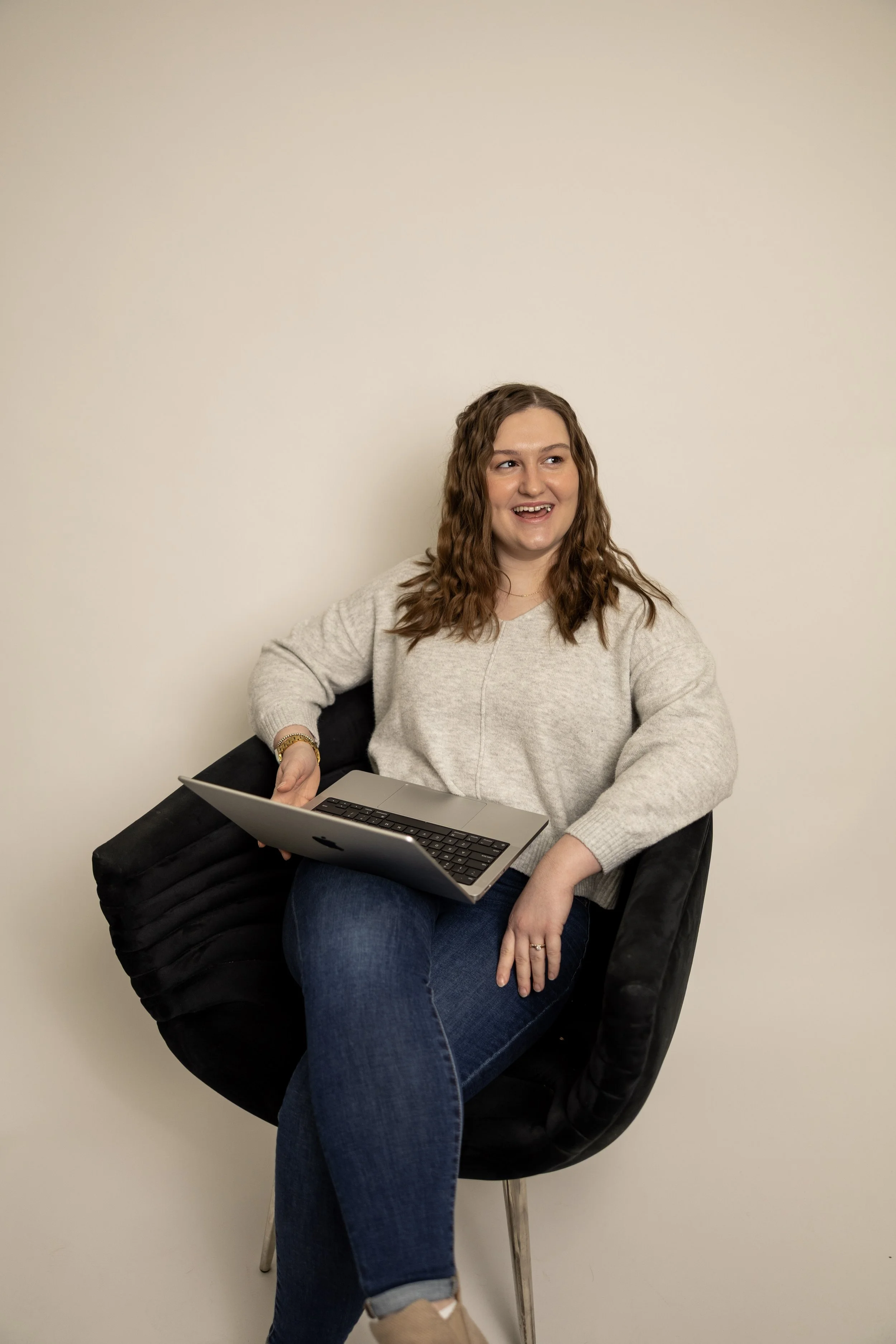 A woman with curly brown hair sitting on a black chair, holding a silver laptop on her lap, smiling and looking to her right, against a plain beige wall.