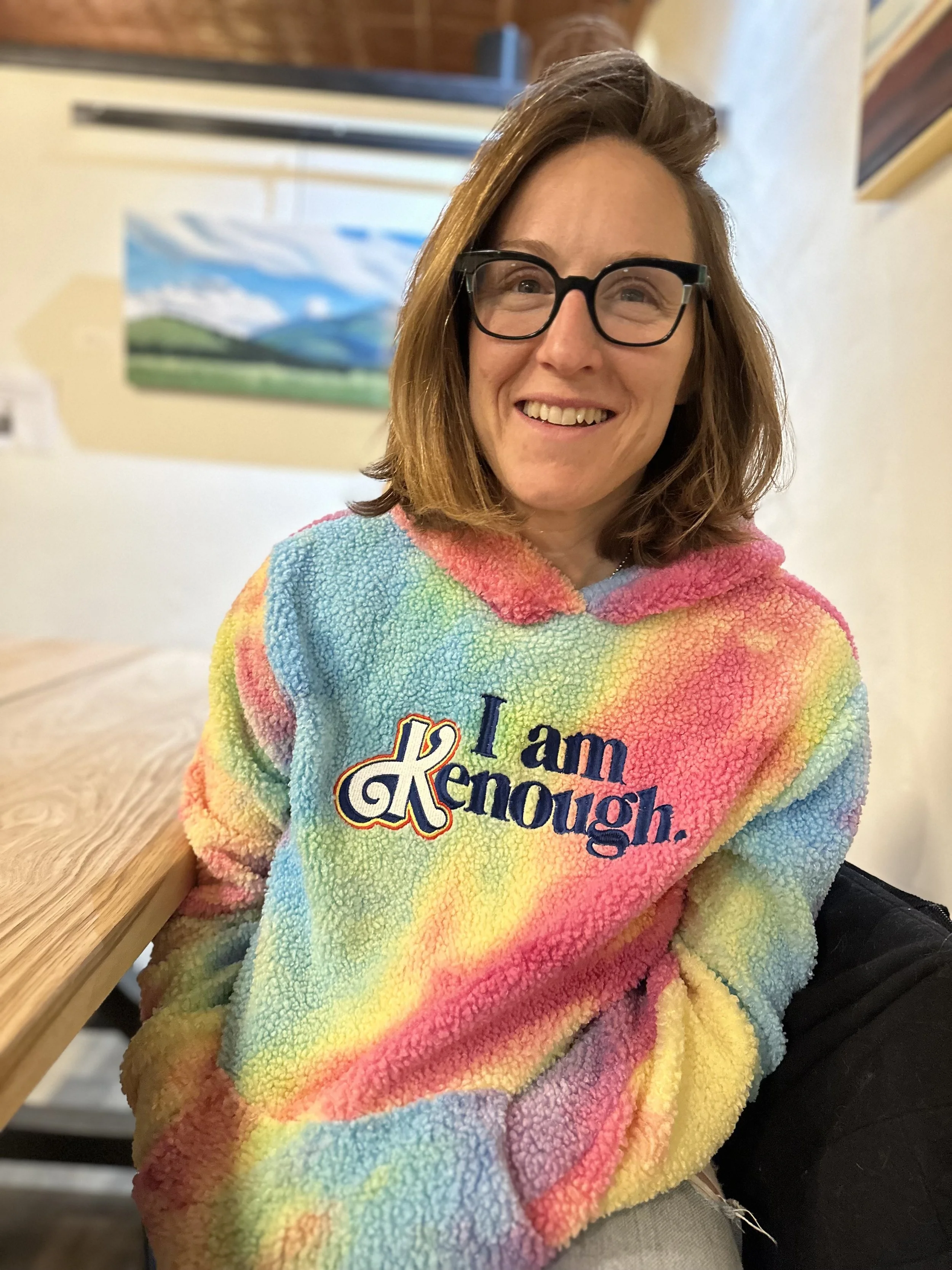 A woman with shoulder-length brown hair and glasses, smiling, wearing a colorful rainbow tie-dye fleece hoodie with the words 'I am kind enough' embroidered on it, sitting at a wooden table in a cozy indoor space.