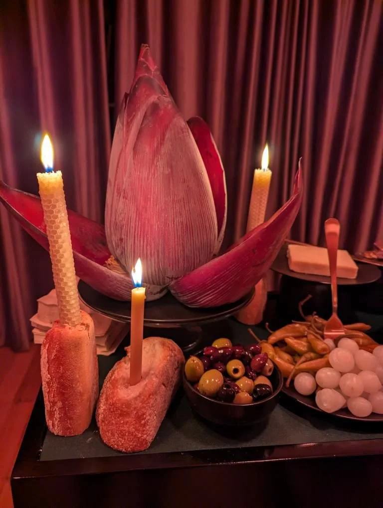 Decorative arrangement with lit candles, large pinkish-red flower petals, bowls of assorted olives, and a plate of white onions, set against a backdrop of red curtains.