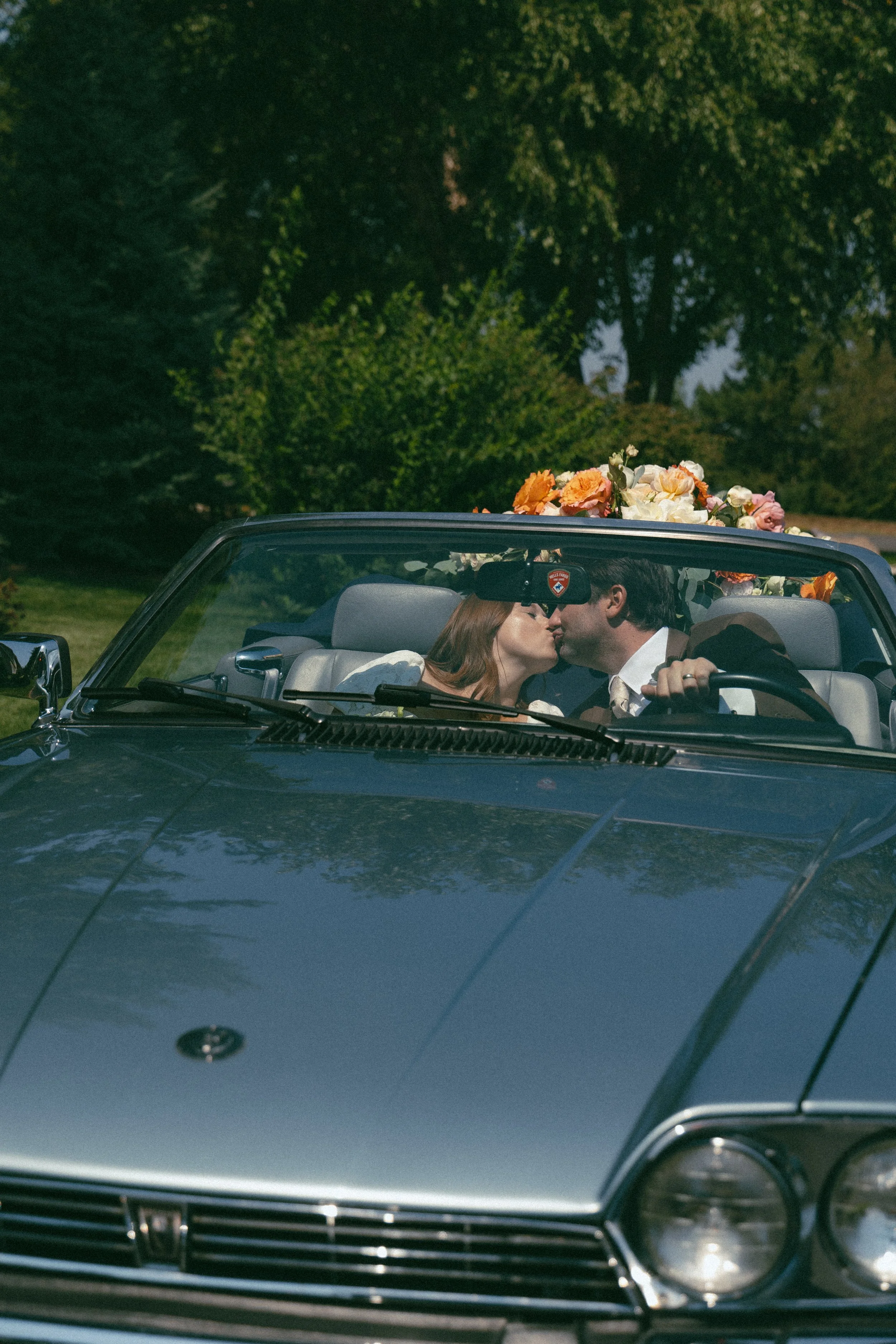 A couple in wedding attire sharing a kiss in a vintage convertible car decorated with flowers.