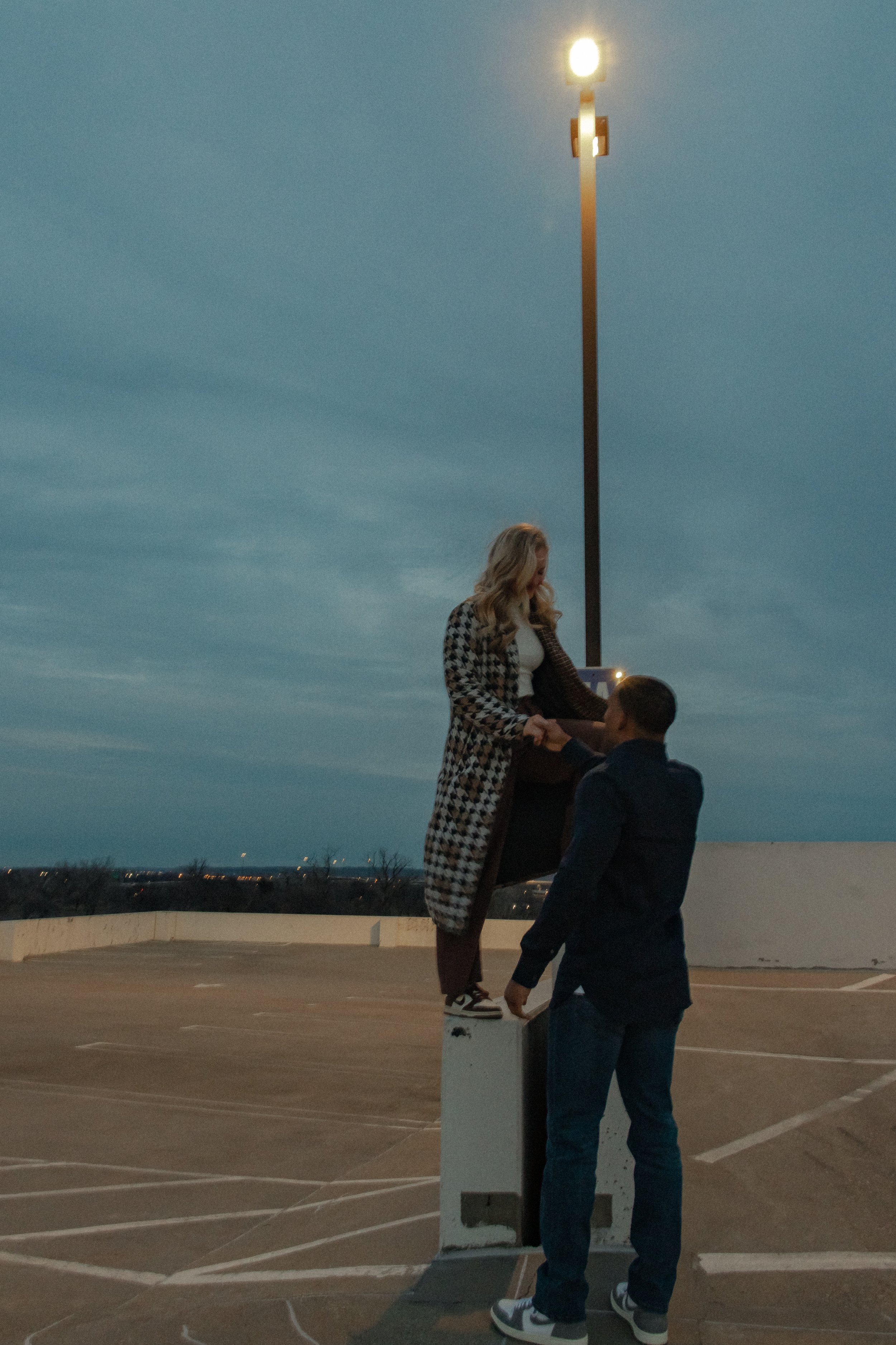 A woman standing on a parking lot barrier holding hands with a man under a tall streetlight with evening sky in the background.