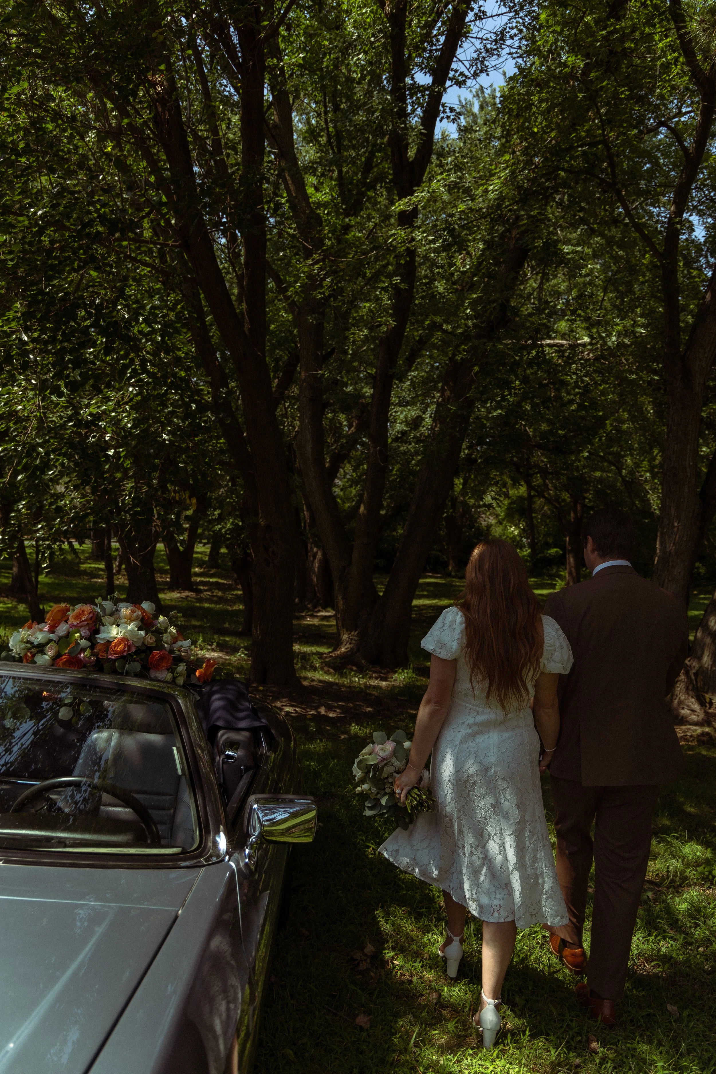 A couple dressed in wedding attire, with the woman in a white lace dress and the man in a dark suit, walking away from a vintage car decorated with flowers in a wooded area.