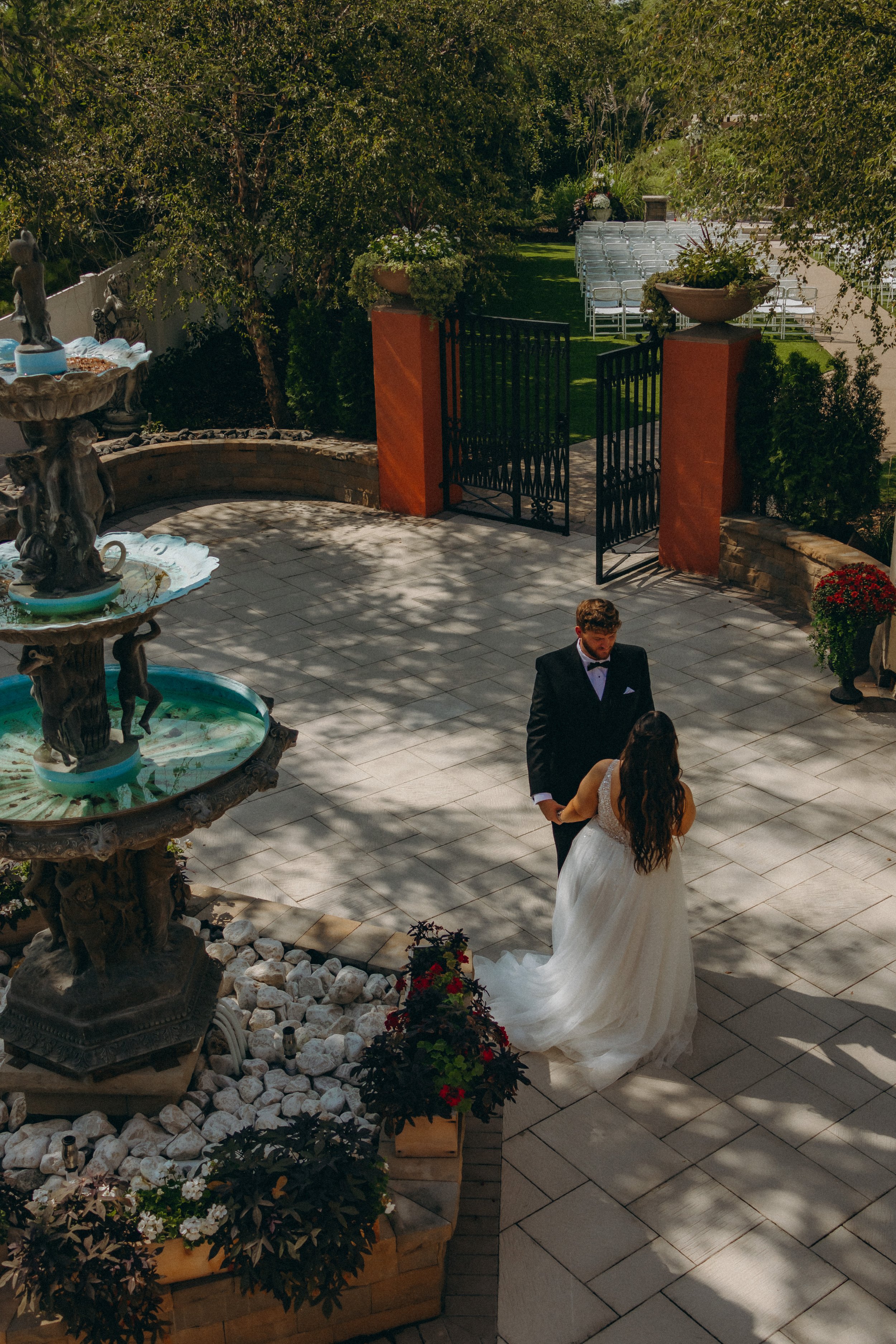 Bride and groom standing near a garden fountain with decorative stone and plants, outdoor wedding setting with chairs in the background.