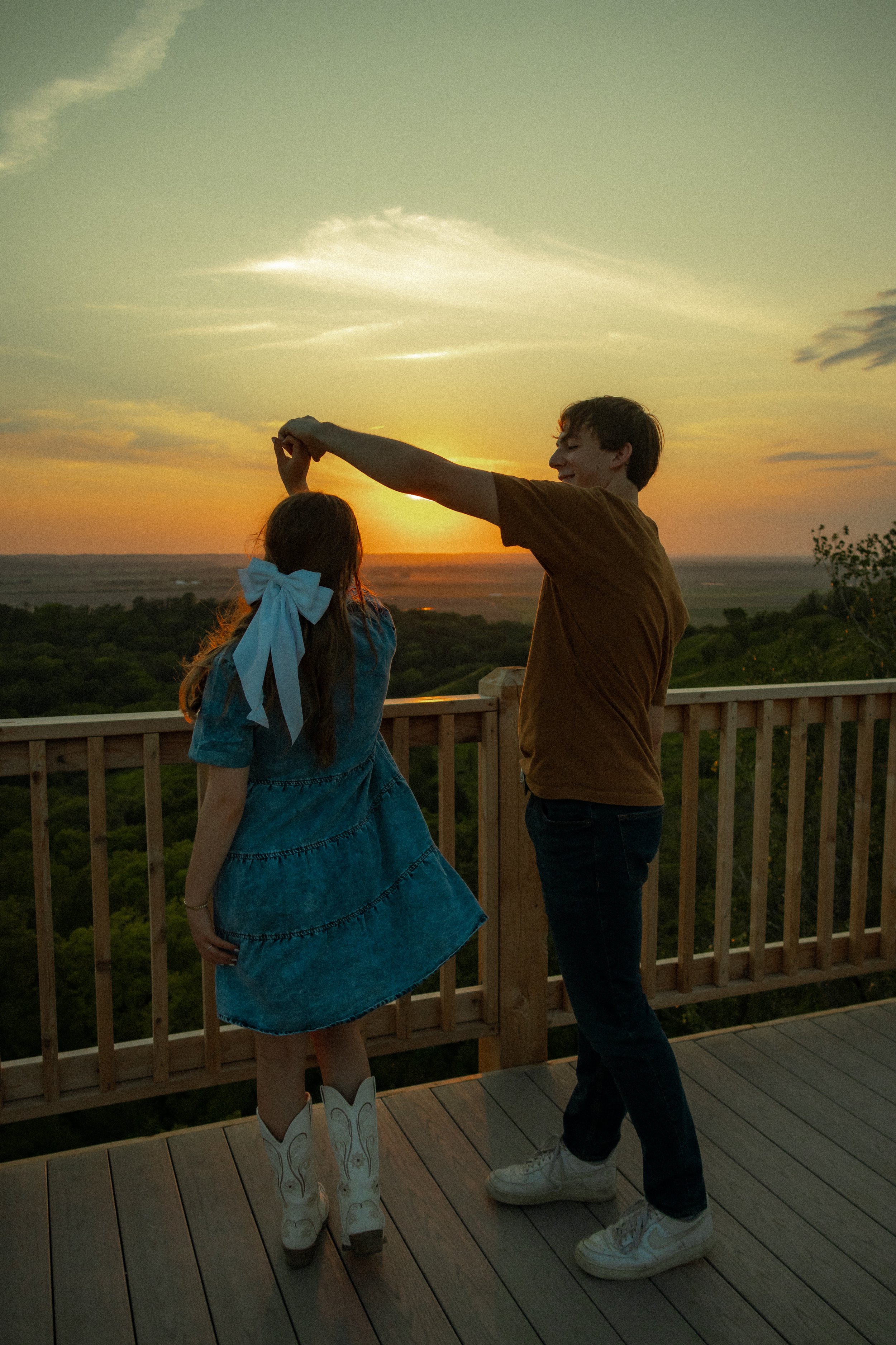 Two people dancing on a wooden deck at sunset, with a scenic view of trees and the horizon.