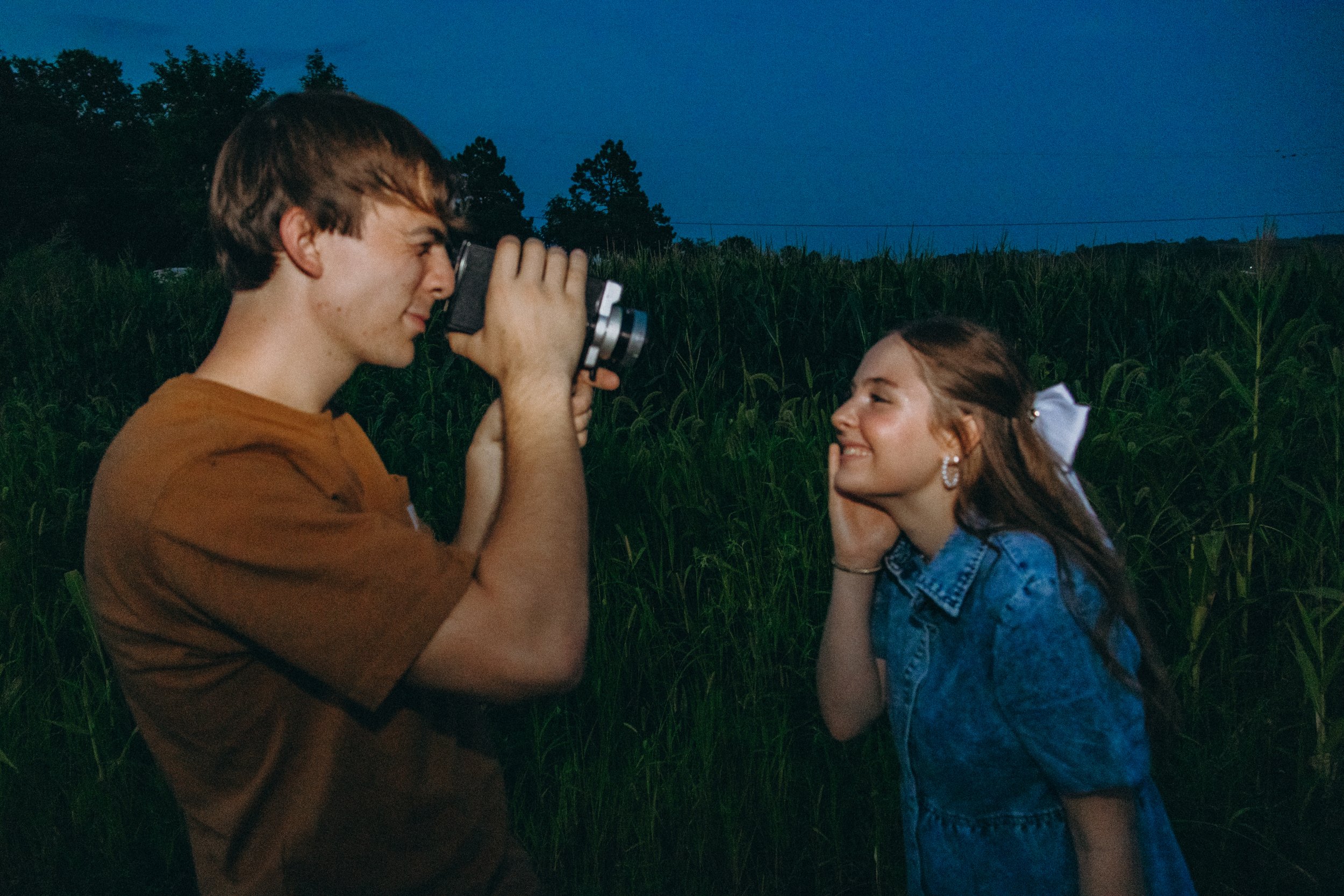 Young man photographing a smiling young woman in a field at dusk.