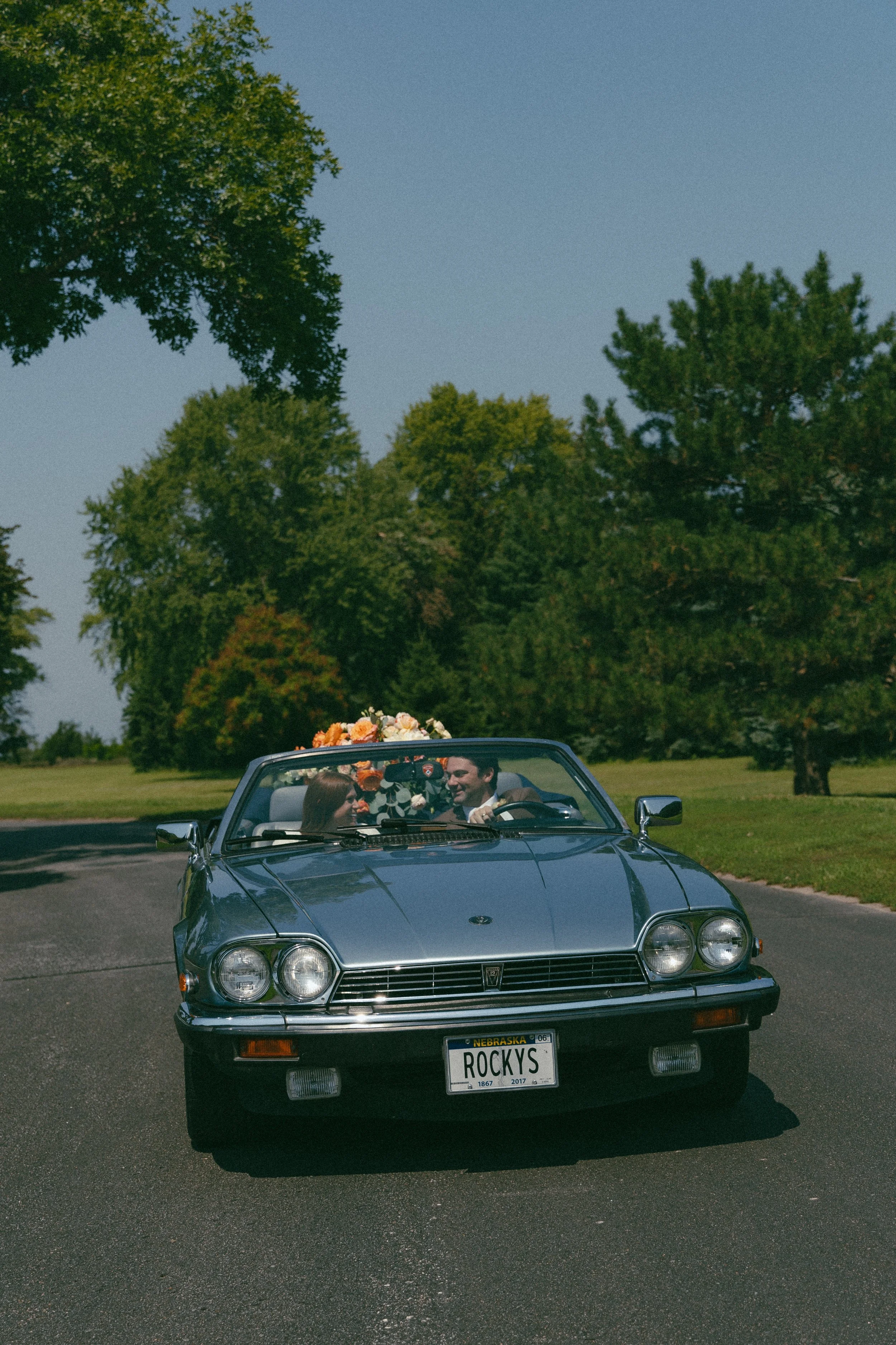 A vintage blue convertible car with a baby on board and a license plate reading 'ROCKYS' is driving on a paved road surrounded by green trees on a sunny day, with two people smiling inside.