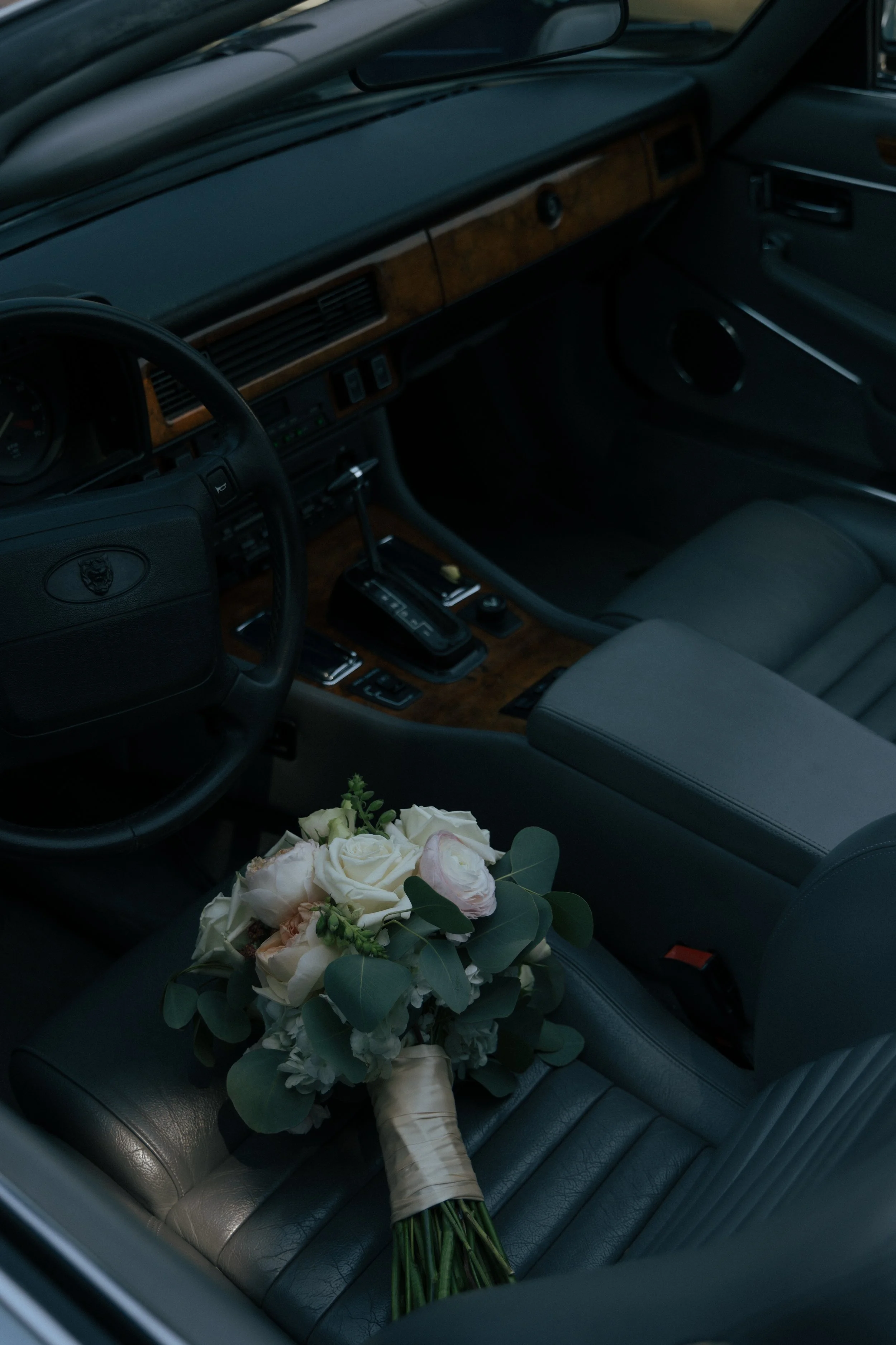 A bouquet of white and pale pink flowers with green leaves resting on the driver's seat of a vintage car interior.