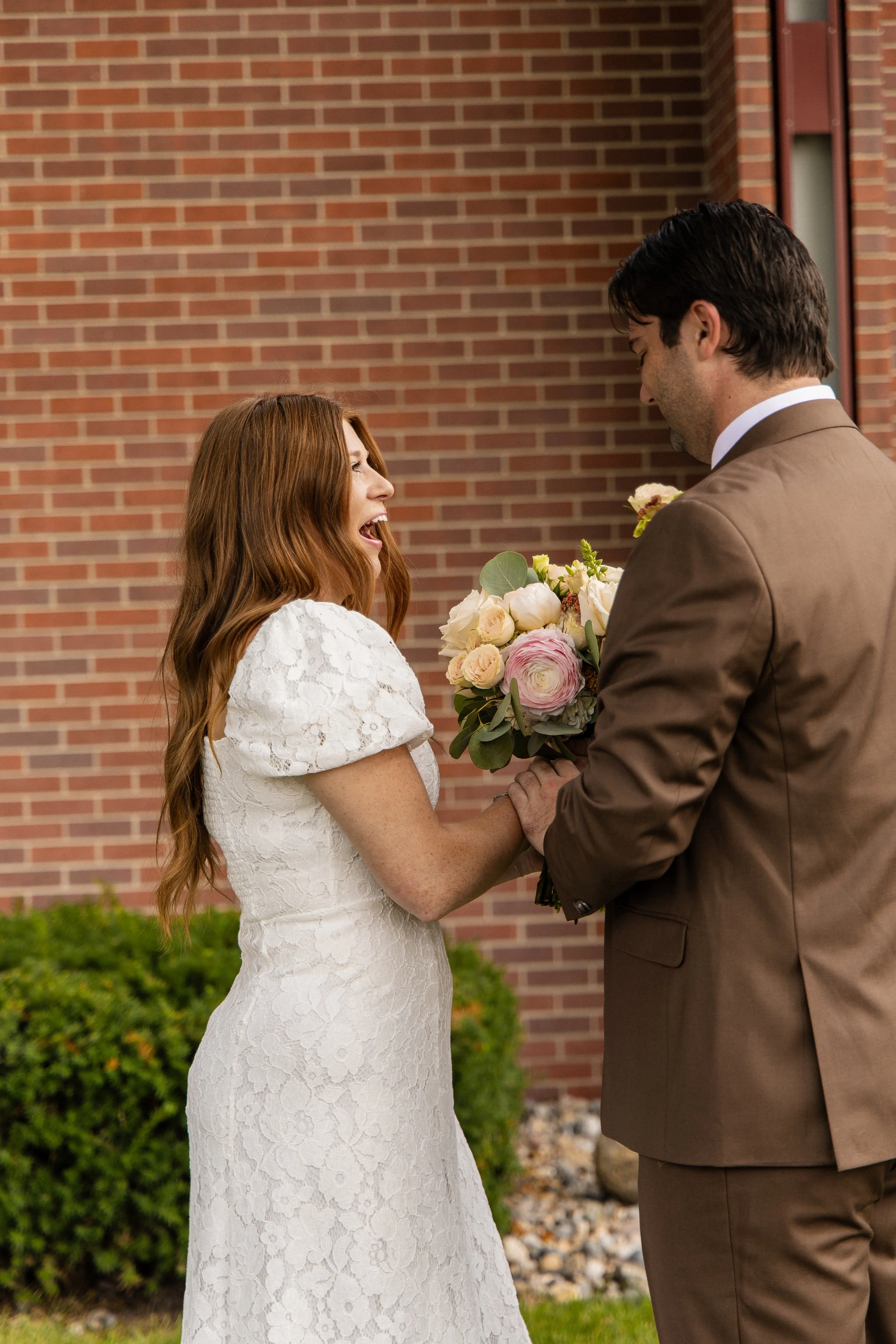 A woman in a white lace dress and a man in a brown suit holding hands and smiling at each other, with the woman holding a bouquet of flowers, against a brick wall background.
