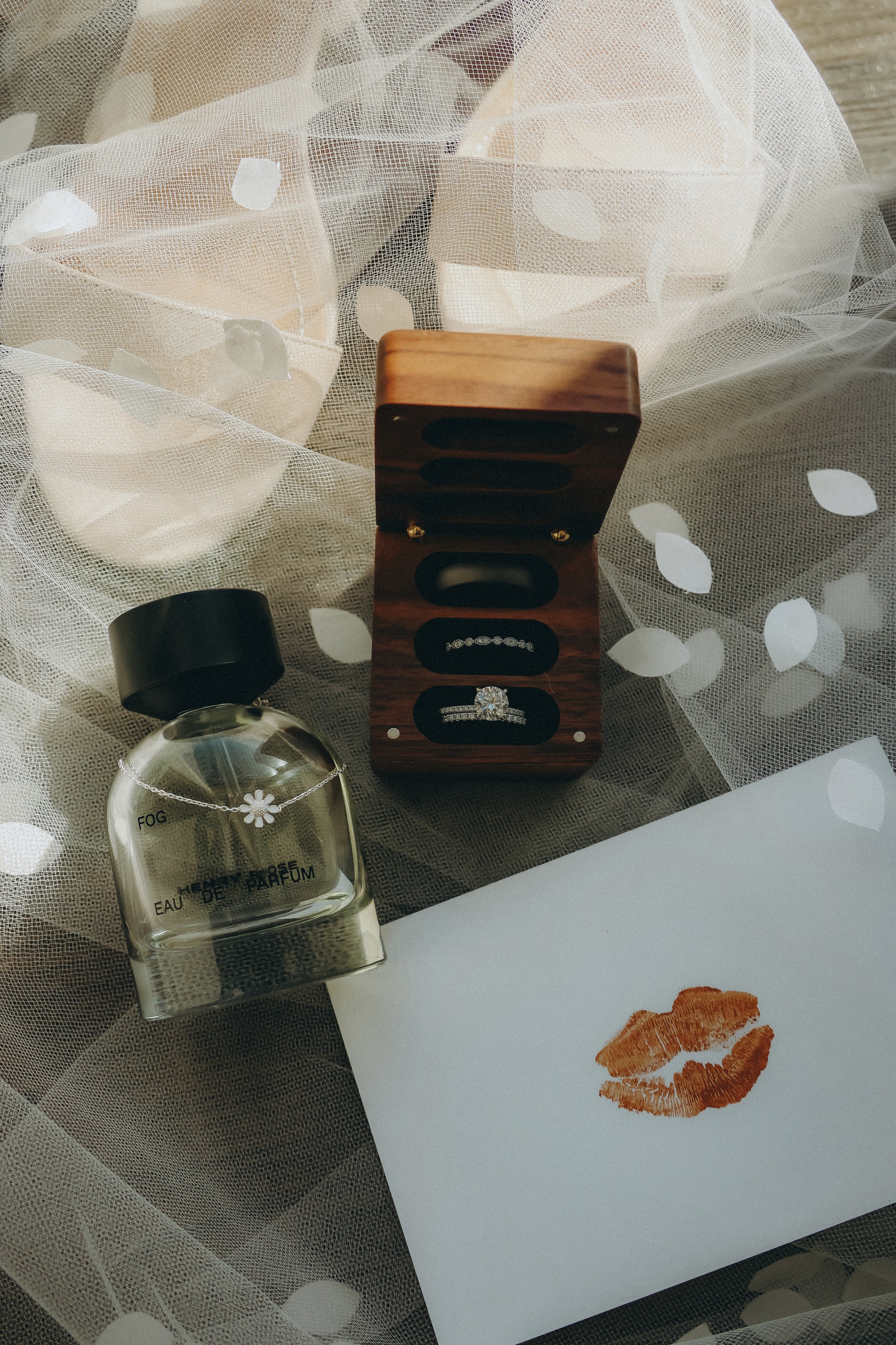 Perfume bottle, open ring box with three rings, and white card with lipstick kiss on tulle fabric.