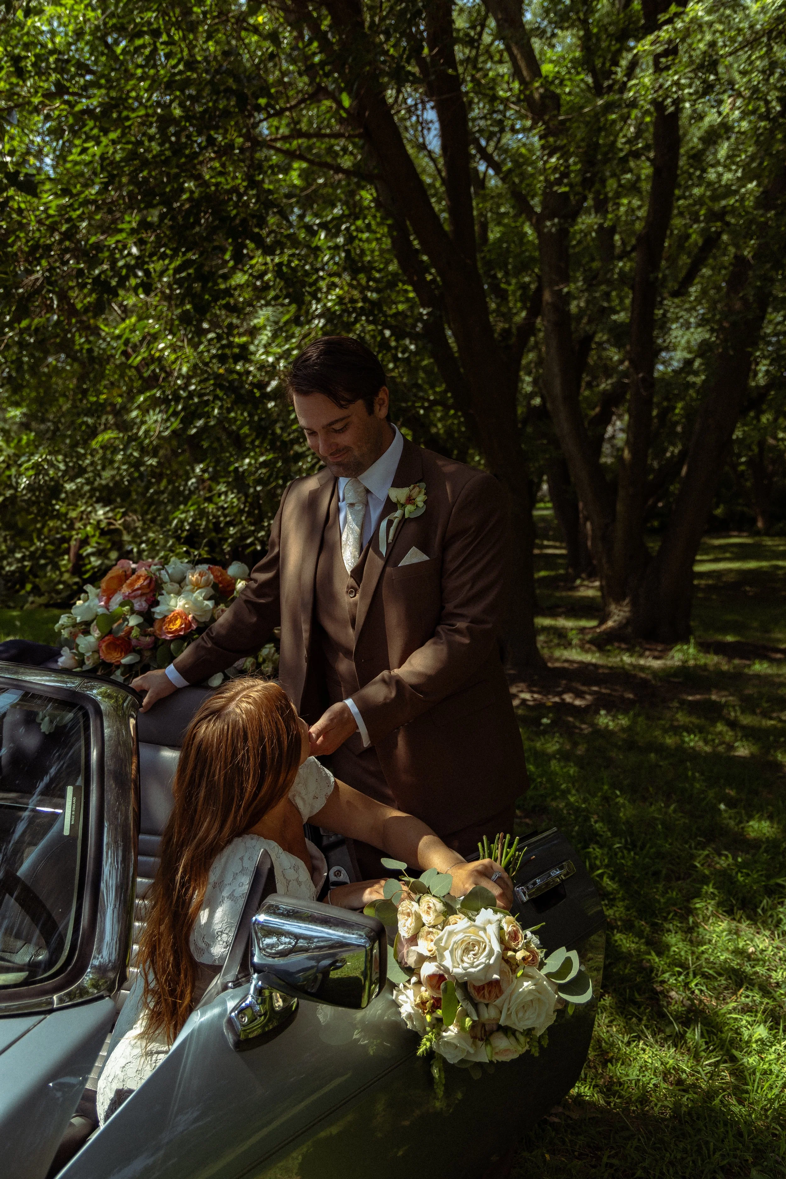 A bride with long red hair sitting in a vintage convertible car, holding a bouquet of white roses and greenery, while a groom in a brown suit with a boutonniere stands outside the car, in a lush green park with large trees.