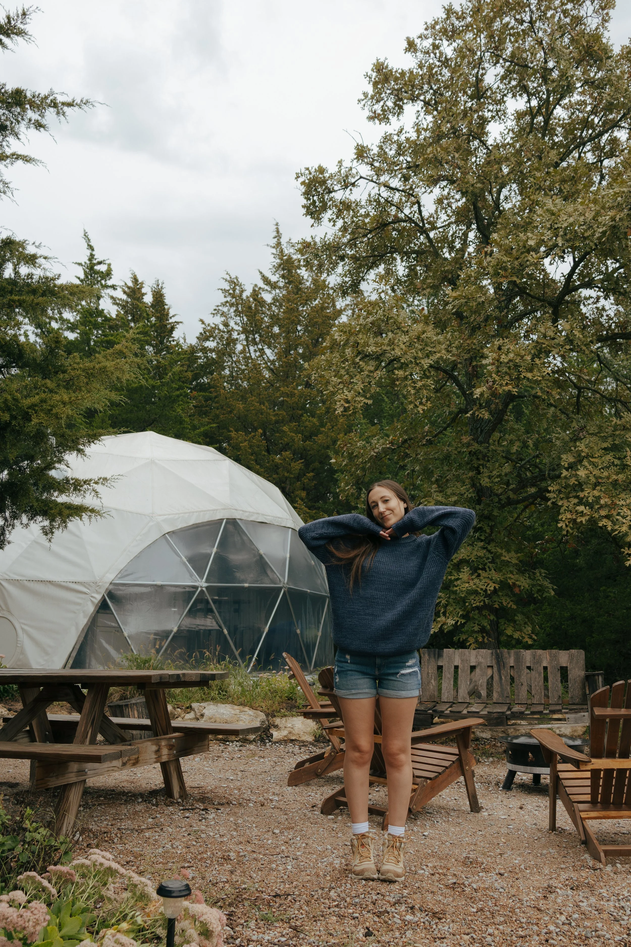 Young woman standing outdoors near a tent and wooden chairs, smiling with arms behind head, surrounded by trees and outdoor furniture.