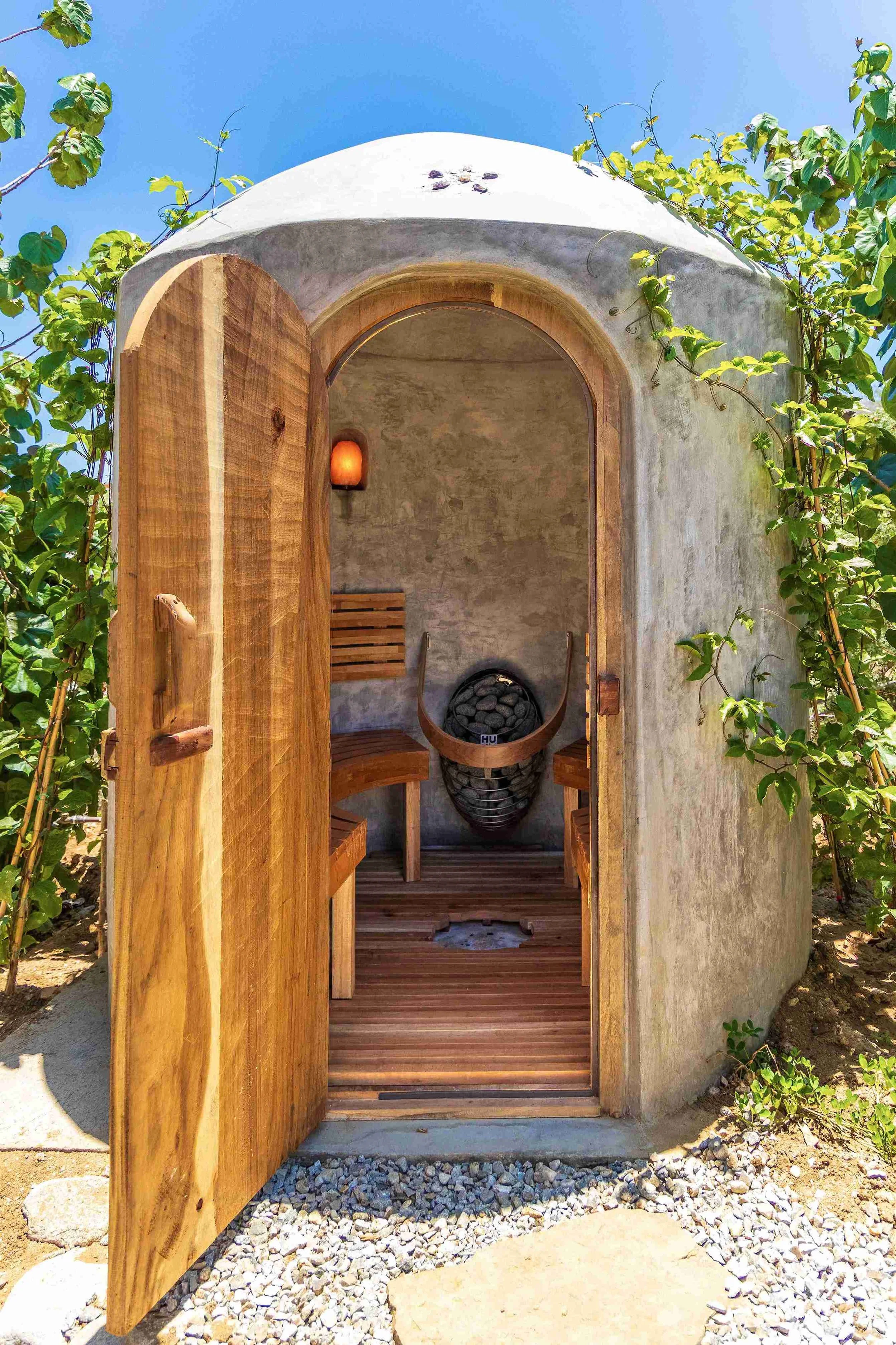Small outdoor sauna or shower with wooden door open, inside a cone-shaped concrete structure, with a chair and fire basket inside, surrounded by green plants and gravel path