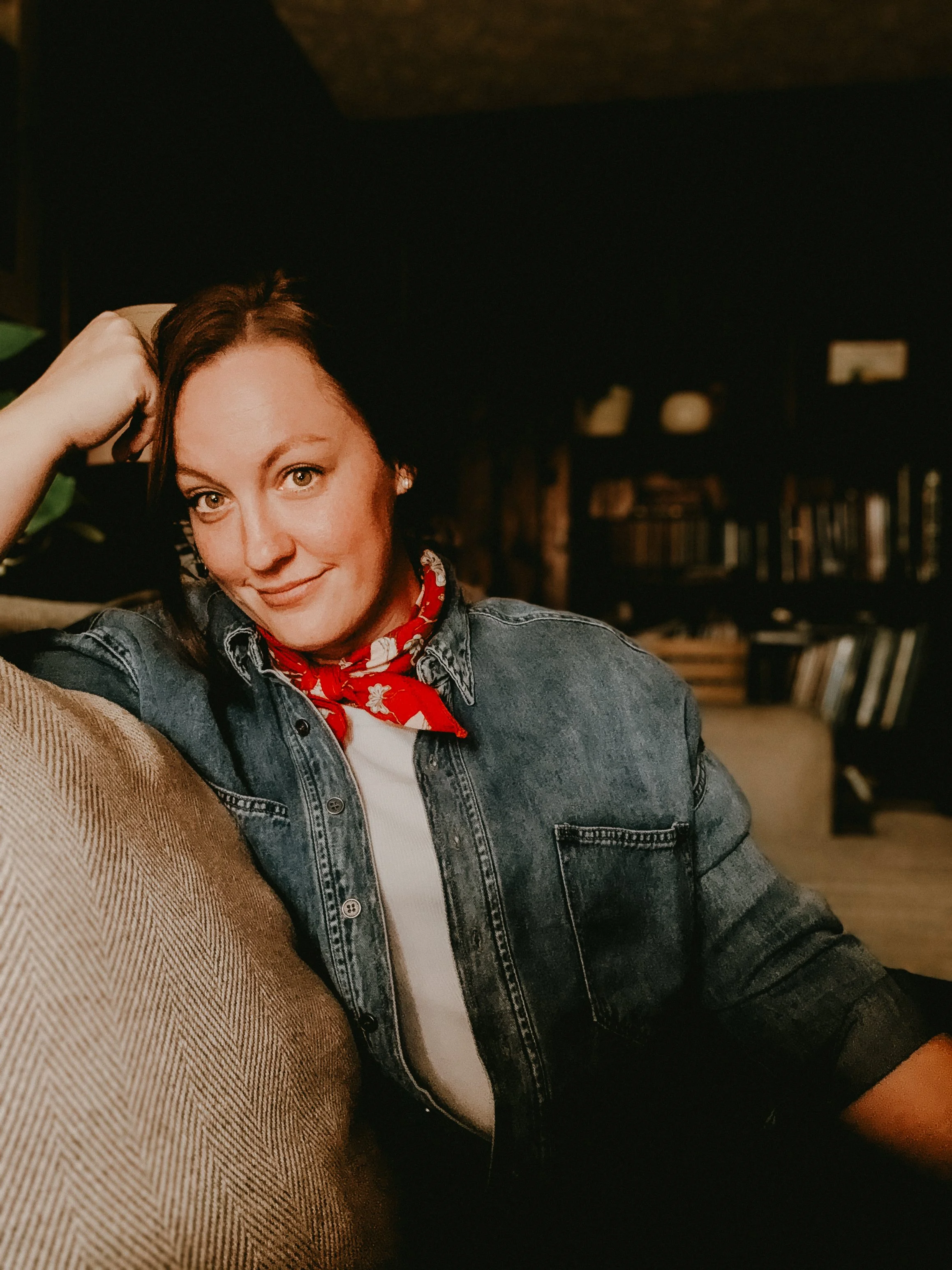 A woman with light skin and brown hair, wearing a denim jacket and a red bandana around her neck, smiling and sitting on a couch in a cozy room with bookshelves in the background.
