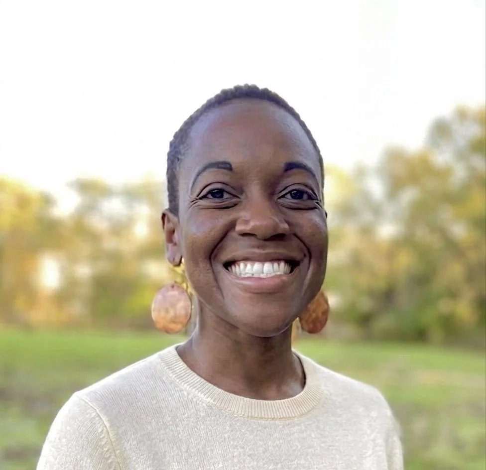 A woman with short hair smiling outdoors in a park during daytime.