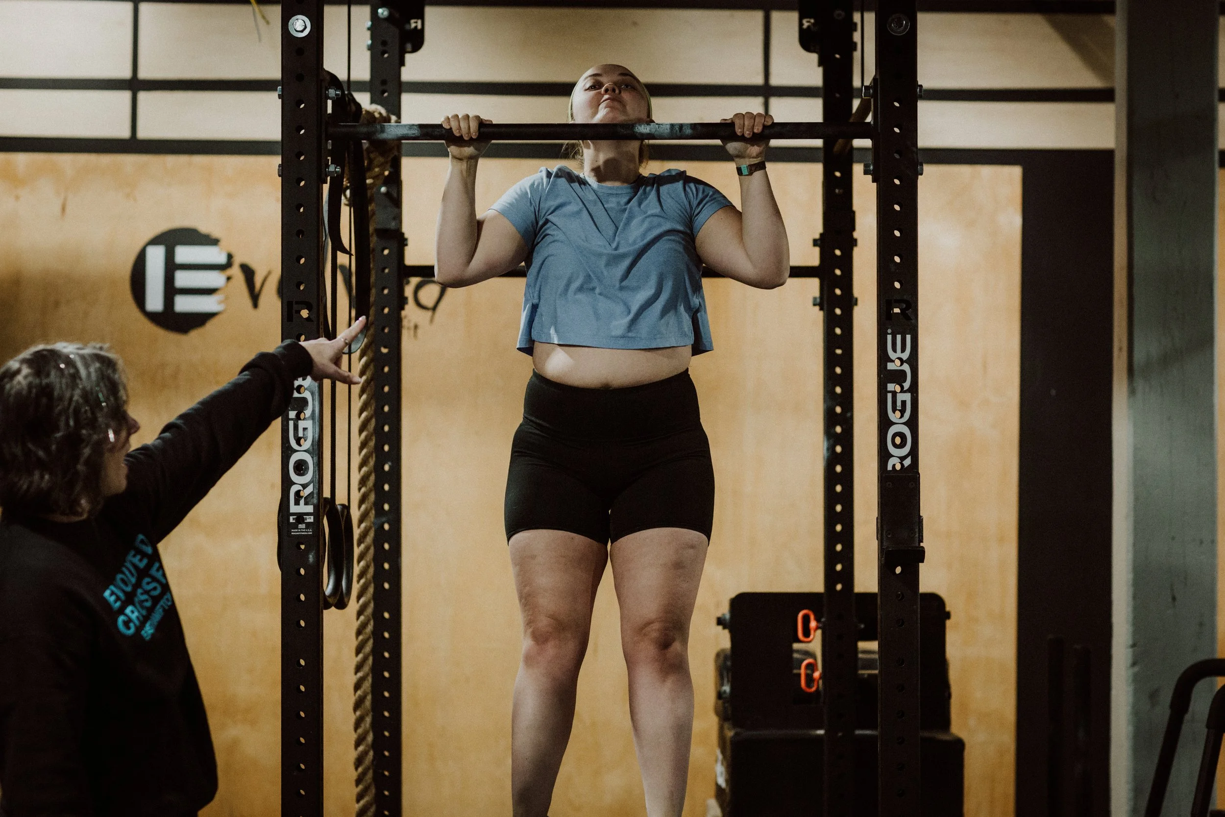 A woman doing a pull-up exercise in a gym, with a trainer pointing at her and giving instructions.
