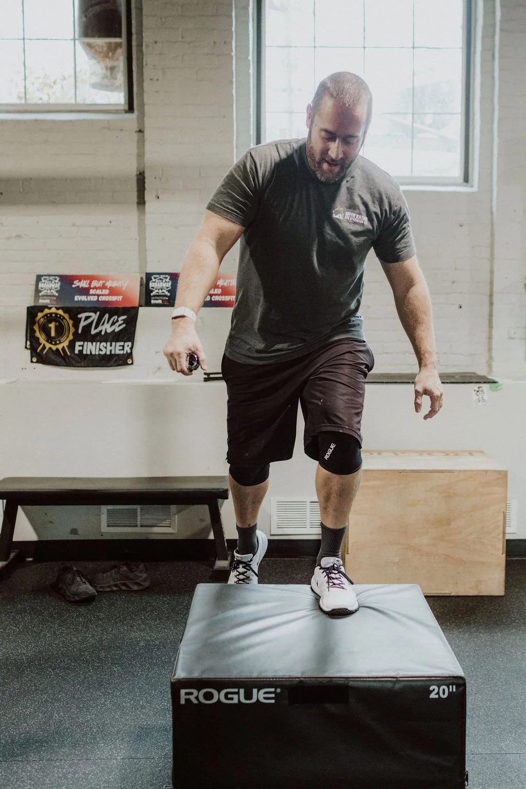Man performing a box jump during a workout in a gym with a "1st Place Finisher" banner and CrossFit signage in the background.