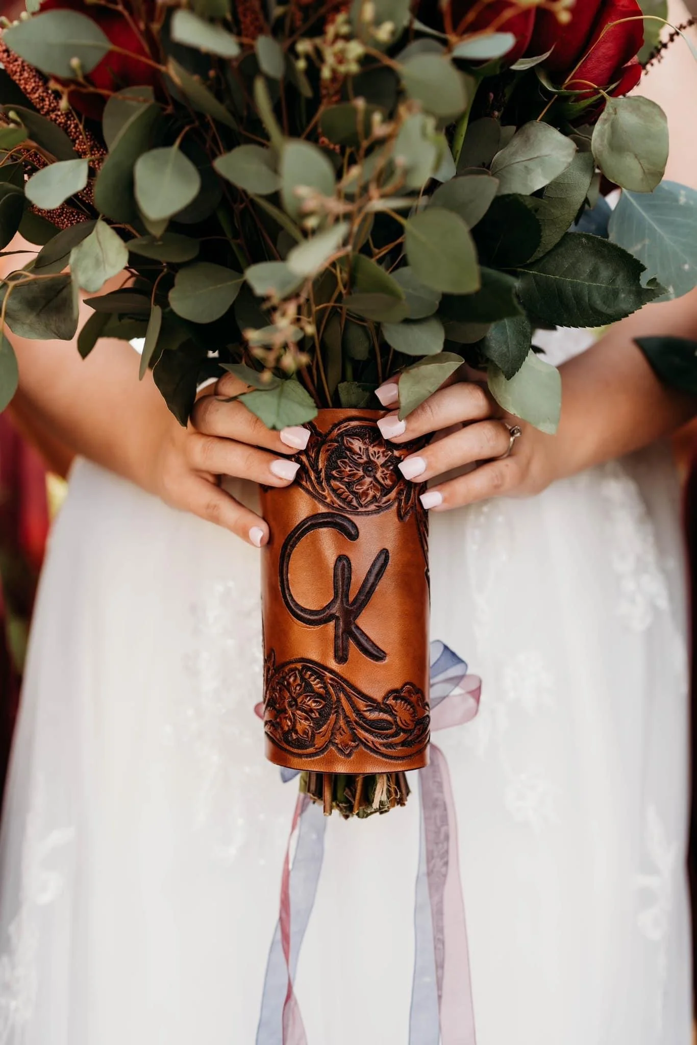Bride holding a bouquet of greenery in a brown leather wrap with initials 'C' and 'K' embossed on it.