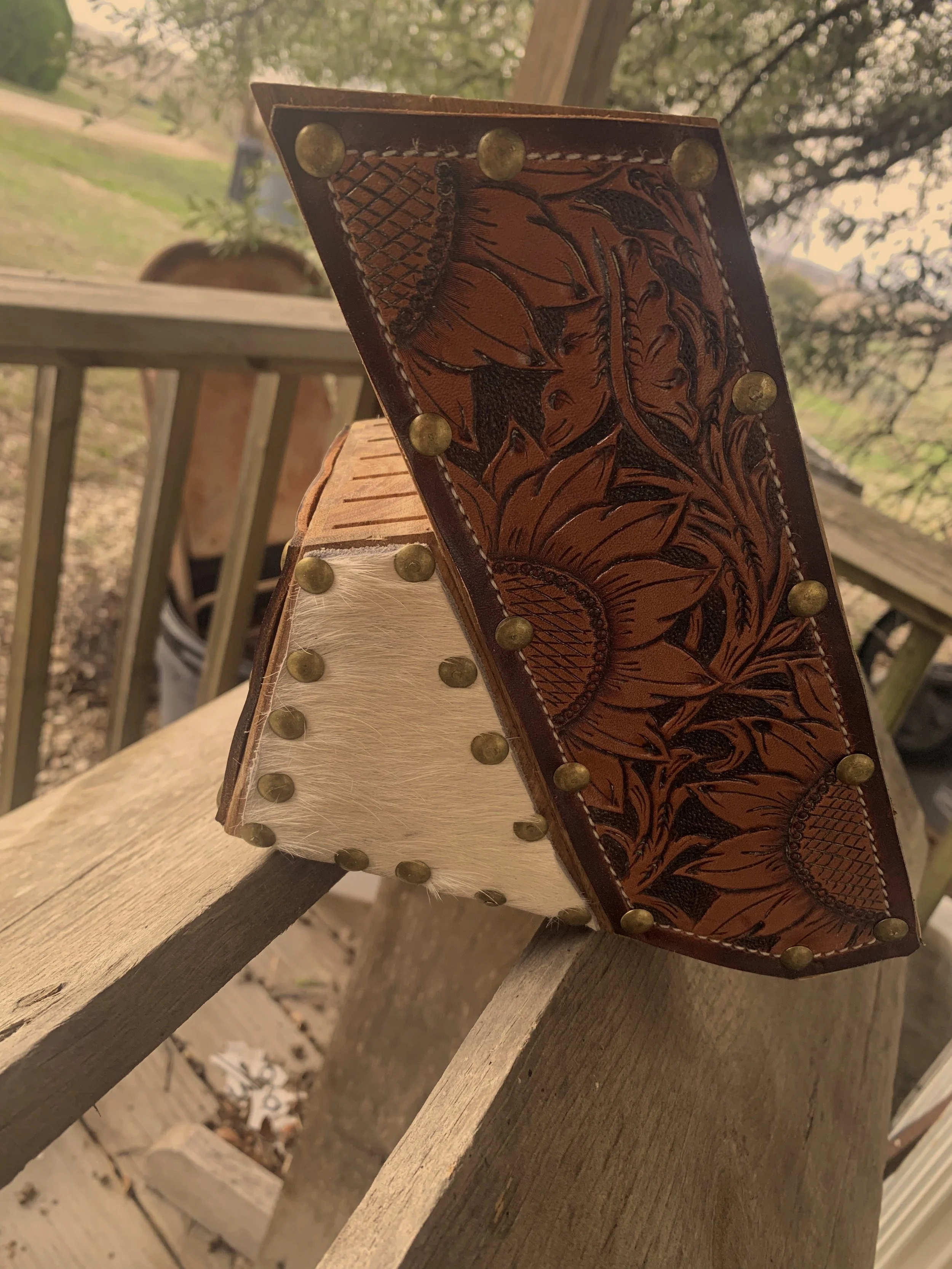 A close-up of a decorative piece with leather, floral, and animal print patterns, attached with brass studs, leaning on a wooden deck railing outdoors.