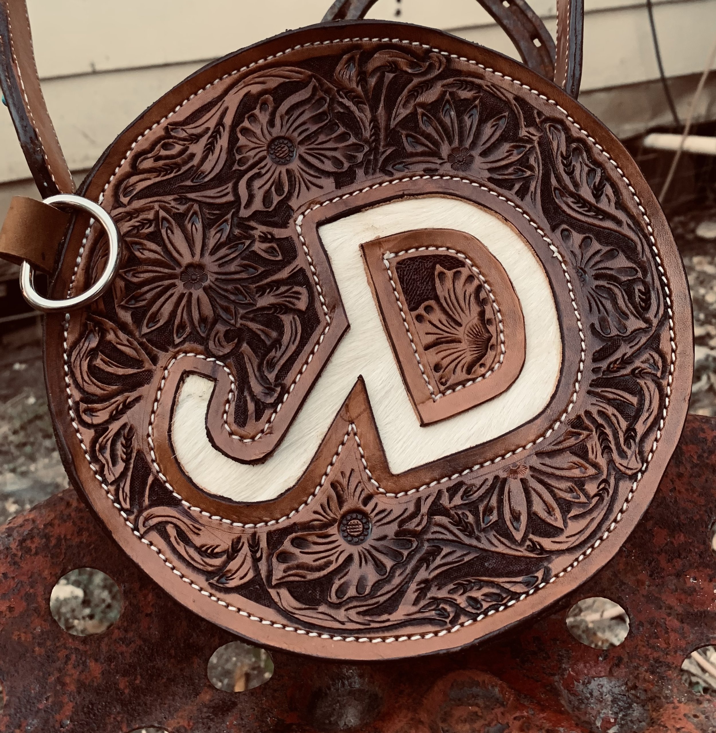 Close-up photo of a round, intricately tooled leather bag or hide featuring a large letter 'D' in the center, with floral and leaf patterns surrounding it. The bag has white and brown contrasting stitching and a metal hook on the side.