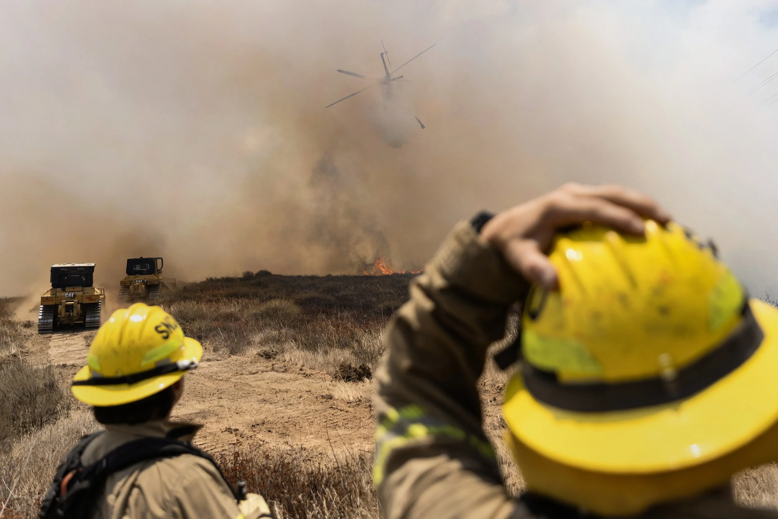 CalFire and San Diego Fire Department crews battle the Sorrento Fire June 8, 2026, in San Diego, Calif. A fire attack helicopter makes a low pass and prepares to drop water on the fire as it begins to crest over the canyon edge. 