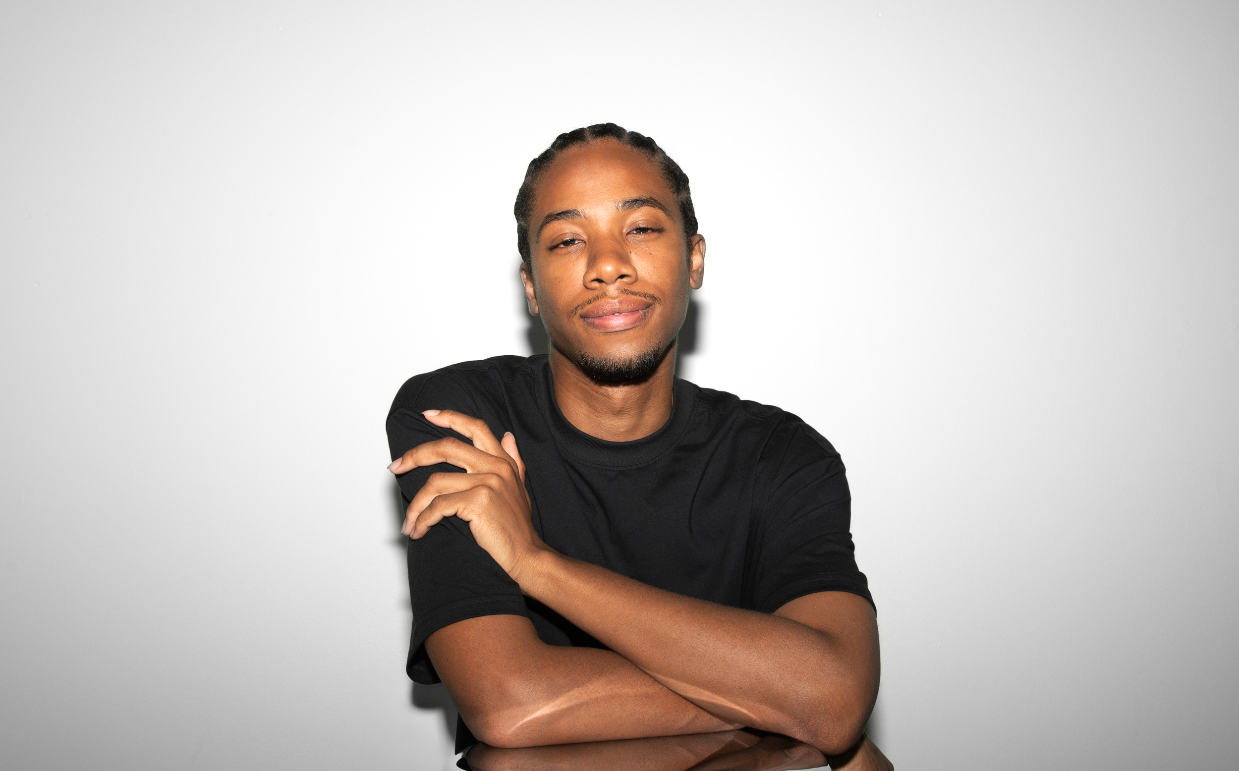 Black, Non-binary person with braided hair, wearing a black t-shirt, standing against a plain white background, smiling with a relaxed expression, crossing his arms.
