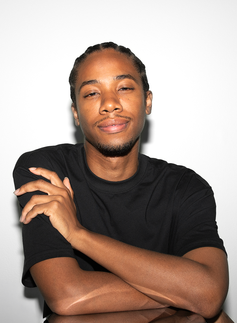 Black, Non-binary person with braids, wearing a black t-shirt, crossing his arms, smiling slightly, against a white background.