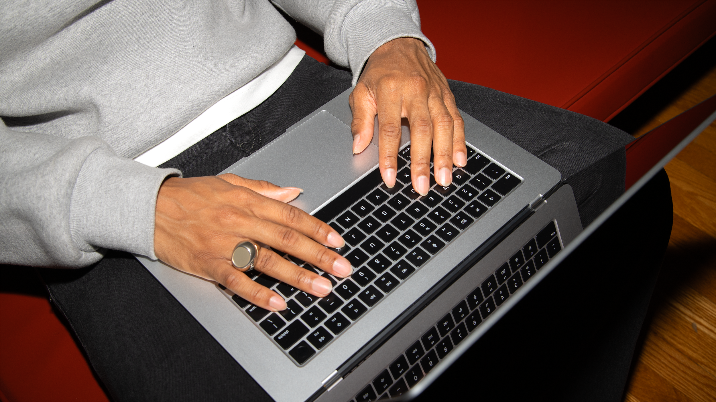 A person wearing a gray sweatshirt and black pants using a silver laptop, with their hands on the keyboard. They have a ring on their left middle finger and a large ring on their right middle finger.