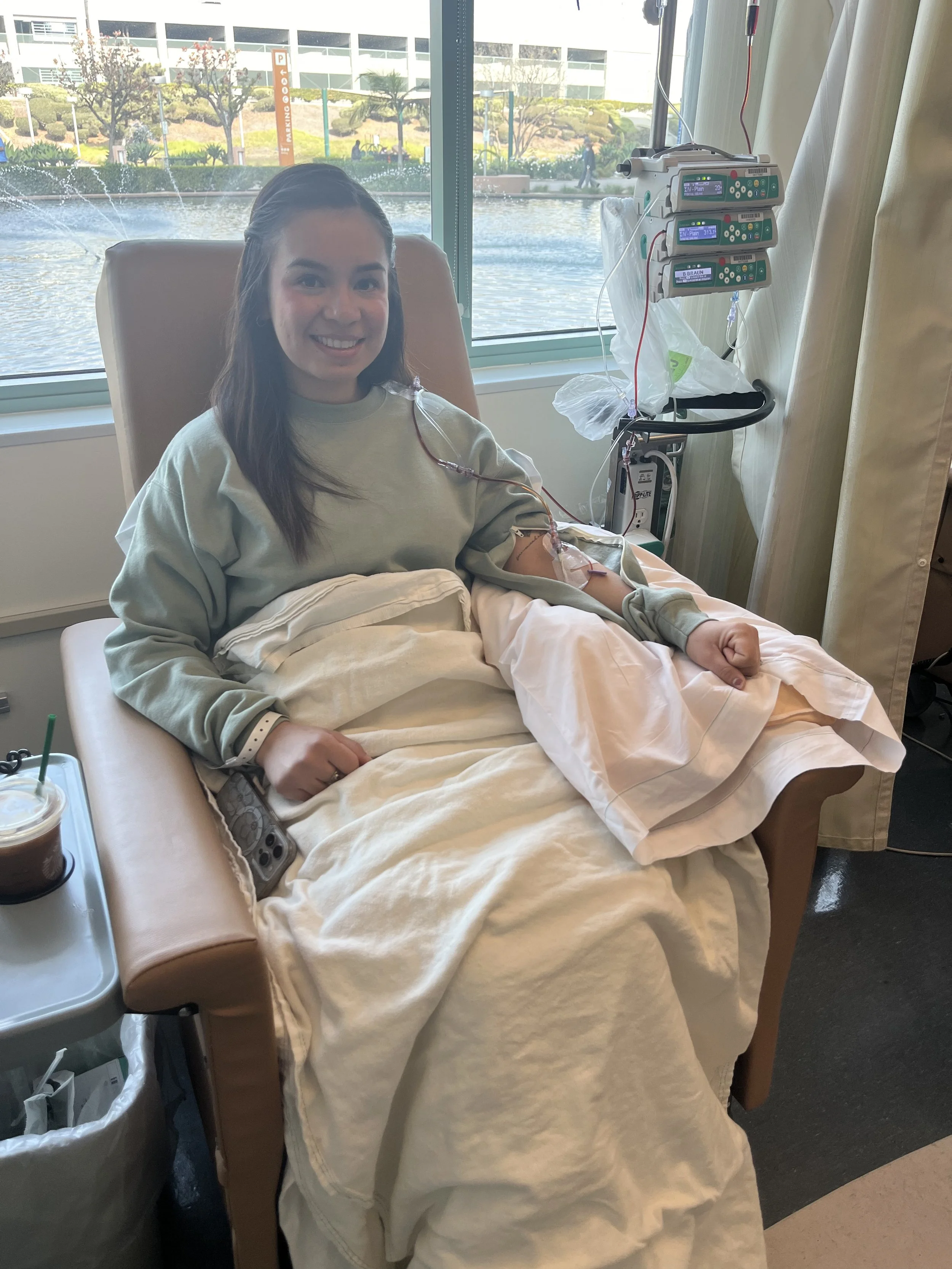A woman seated in a hospital bed, smiling. She has an IV inserted in her arm connected to a monitoring machine, and a tray with a drink on her side. The hospital room has large windows showing a view outside with water and trees.