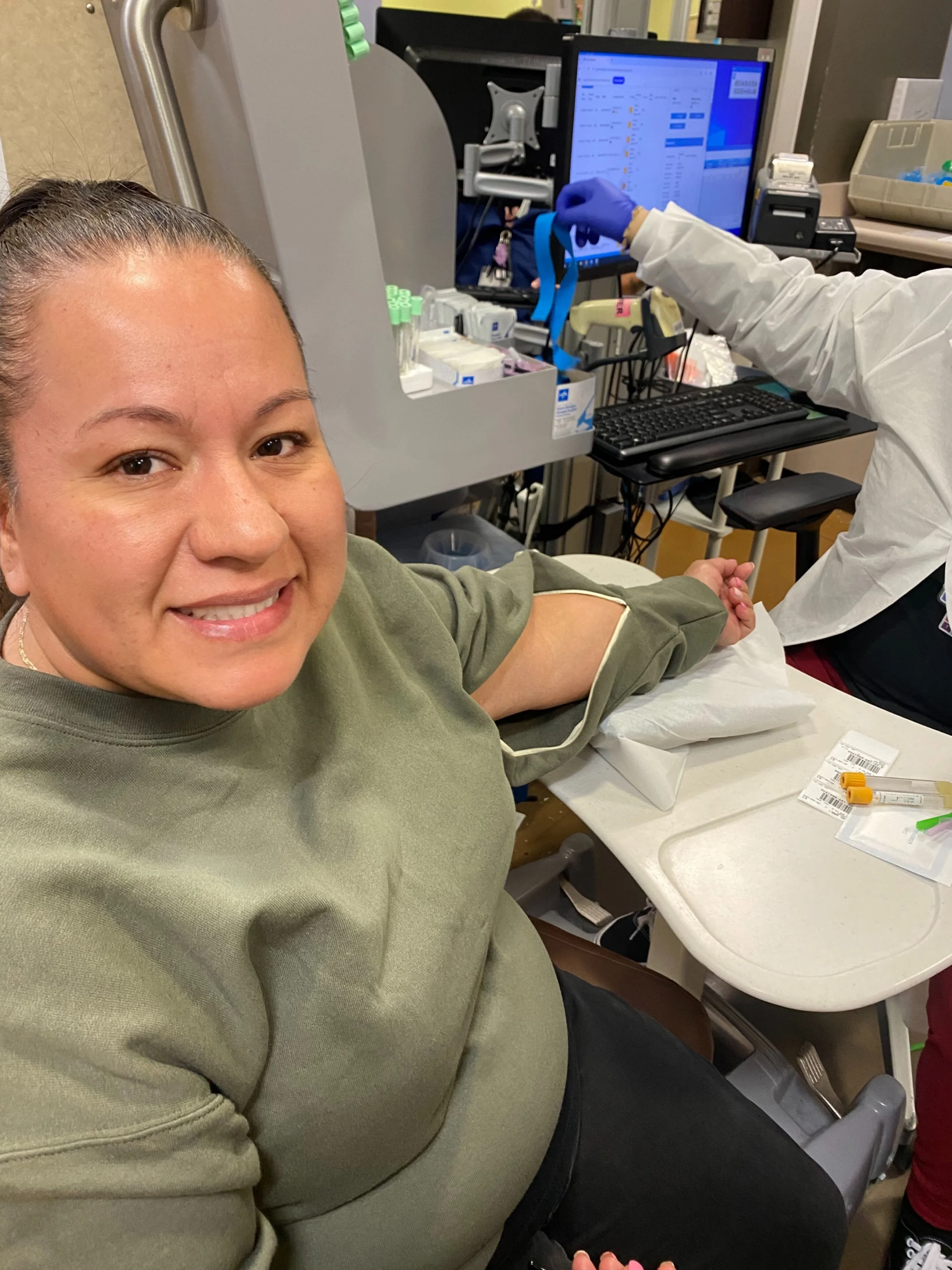 A woman with a smile, sitting in a medical chair, having her arm vaccinated by a healthcare worker in a clinical setting. The healthcare worker is wearing gloves and a white coat, and there is medical equipment and a computer on the desk nearby.