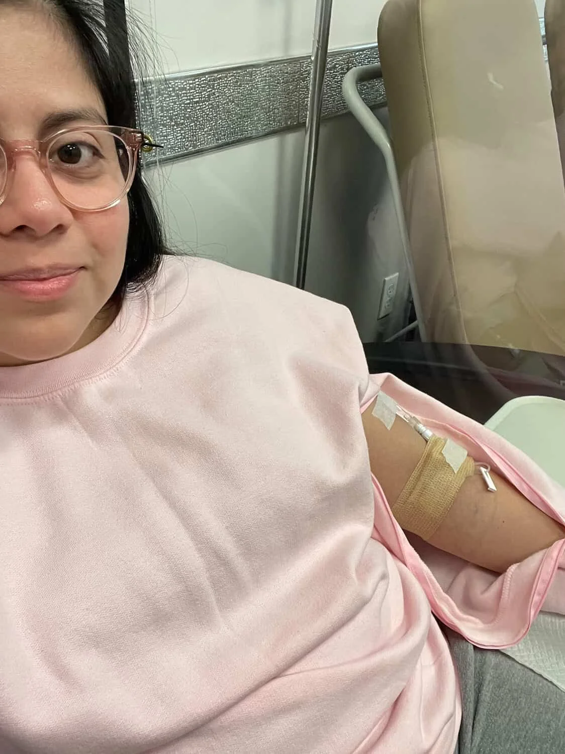 A woman wearing glasses and a pink shirt is sitting in a medical setting, donating blood, with a needle and tube in her arm.
