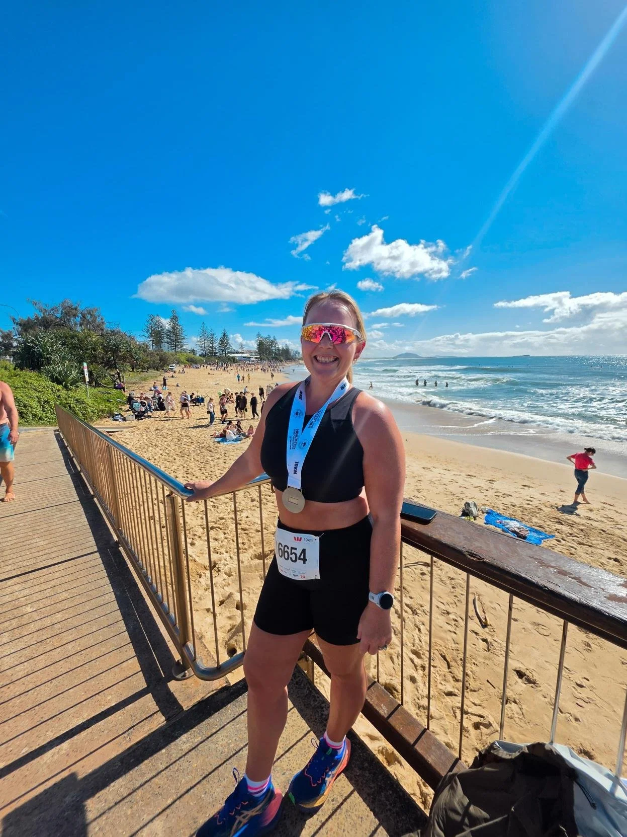 Woman wearing athletic gear with a race bib and medal, standing on a boardwalk at a sunny beach with people, sand, ocean, and blue sky in the background.