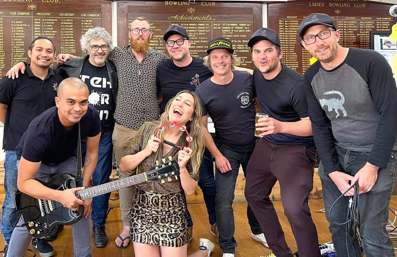 Live band and friends posing together inside Footscray Park Bowling Club, with a guitarist and tambourine player at the front and the club’s honor boards in the background.