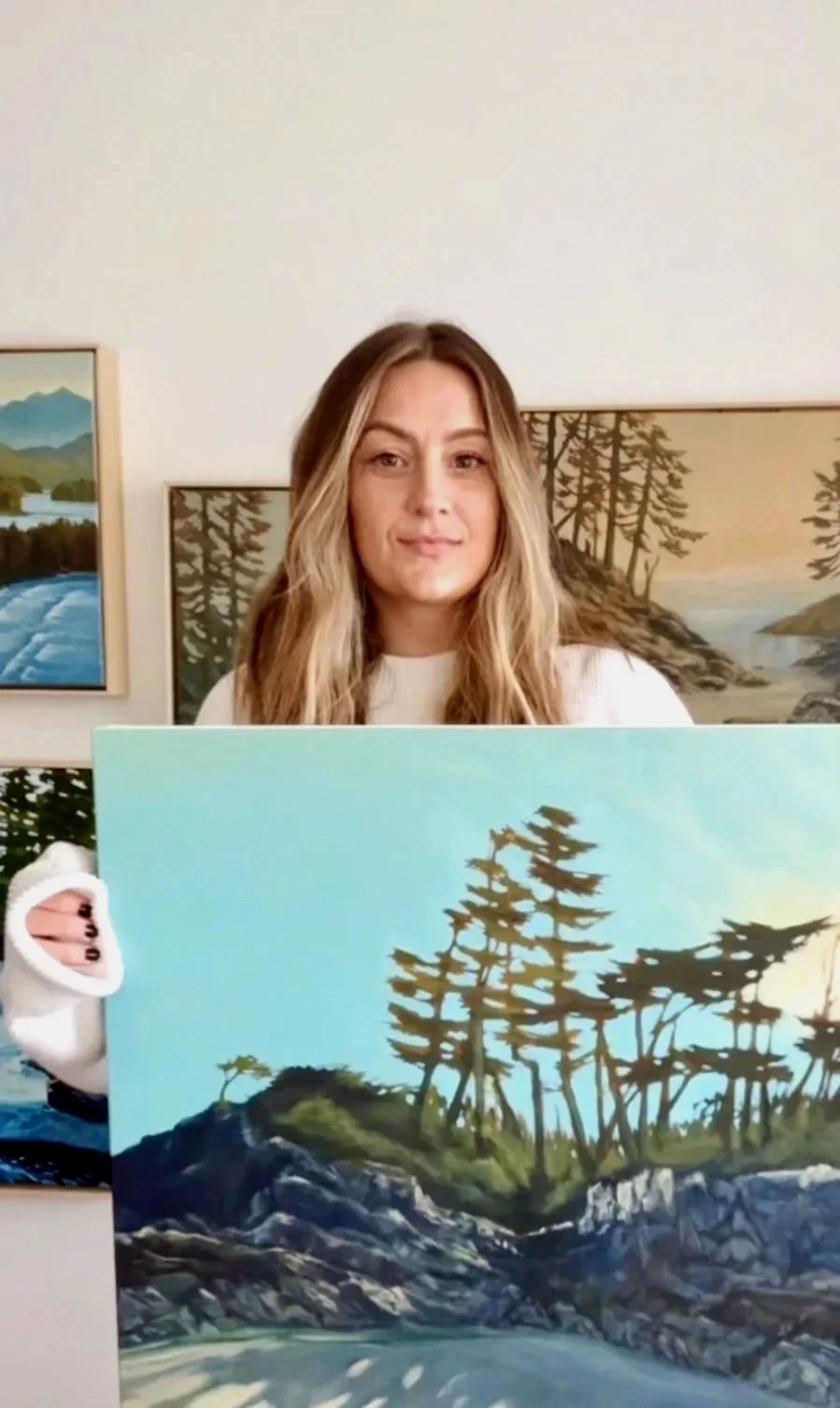 Woman with wavy blonde hair standing indoors, holding a landscape painting of trees and rocks near water, with similar paintings on the wall behind her.