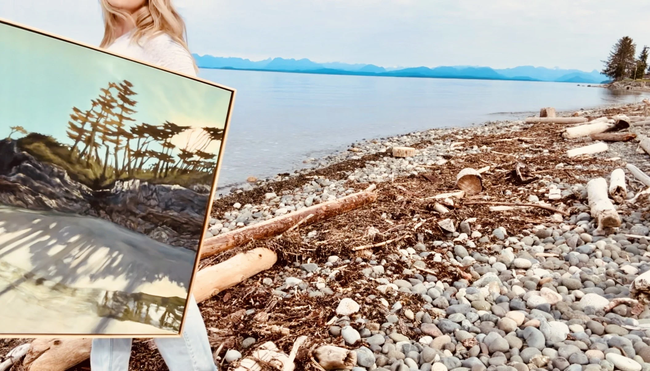 A person holding a framed landscape painting in front of a rocky beach with driftwood, pebbles, and a body of water with mountains in the background