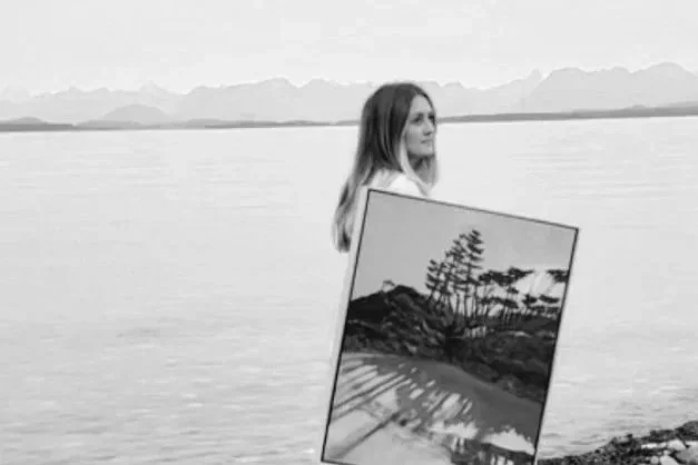 A woman standing by a body of water holding a painting of a landscape with trees and mountains, with mountains visible in the distance.