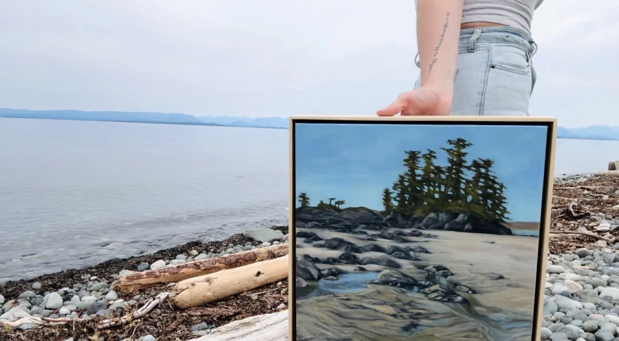 A person holding a framed painting on a rocky beach with driftwood and water in the background, depicting a similar shoreline with trees and water.