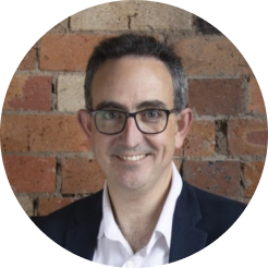 Michael Strachan - man with glasses and blazer smiling in front of brick wall