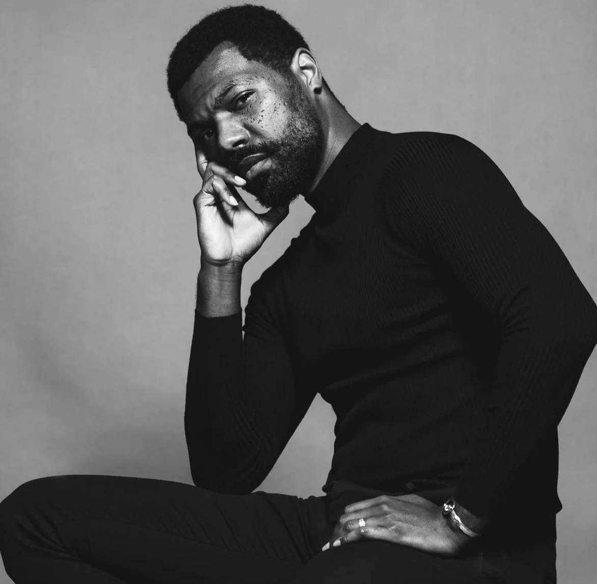 Black and white portrait of a man with a beard, wearing a long-sleeve shirt, sitting with his hand resting on his face, looking at the camera.