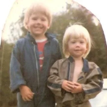 Two young children with blond hair standing outdoors and smiling at the camera.