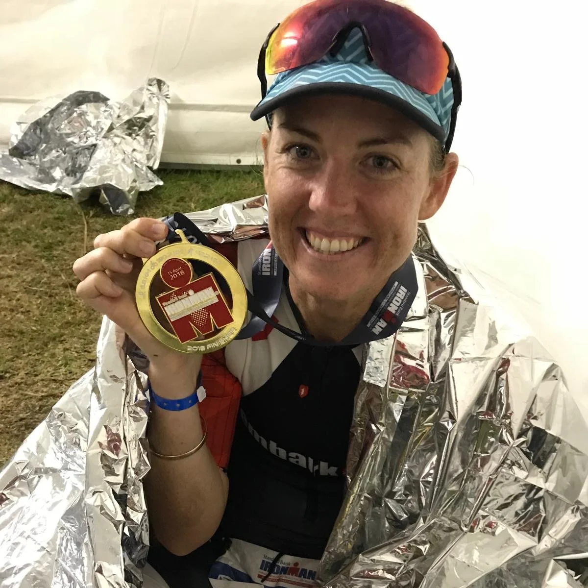 A smiling woman wearing a cap, sunglasses on her head, and a silver foil blanket, holding a finisher medal from Ironman 2018. She is inside a tent or sheltered area.