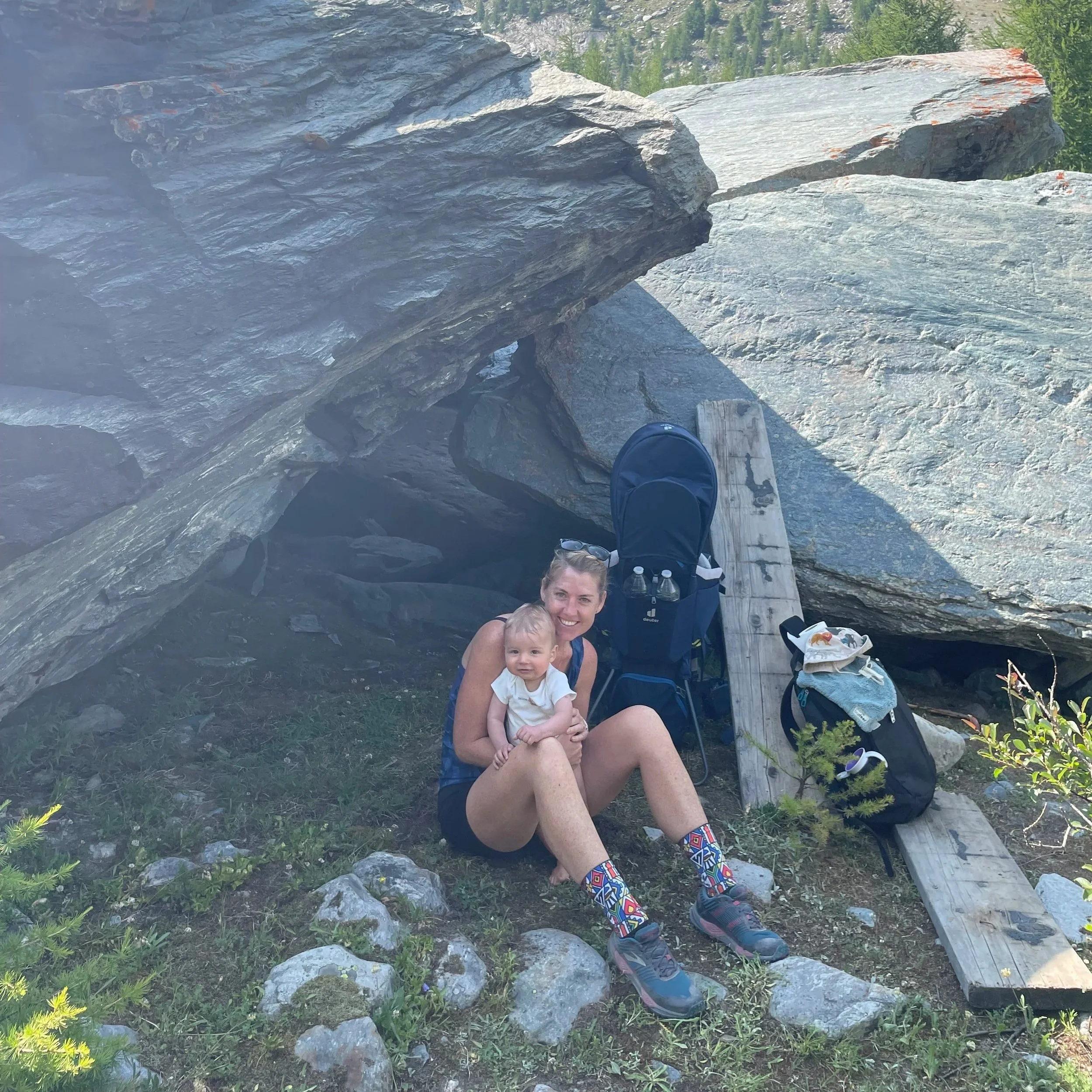 A woman with a baby sitting on rocks in front of a large rock formation outdoors, smiling at the camera, with hiking gear and backpacks nearby.
