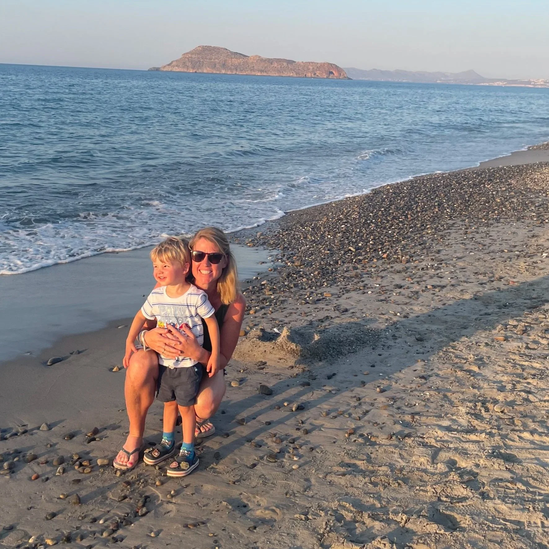 A woman and a young boy smiling on a pebble beach near the water, with an island in the distance.